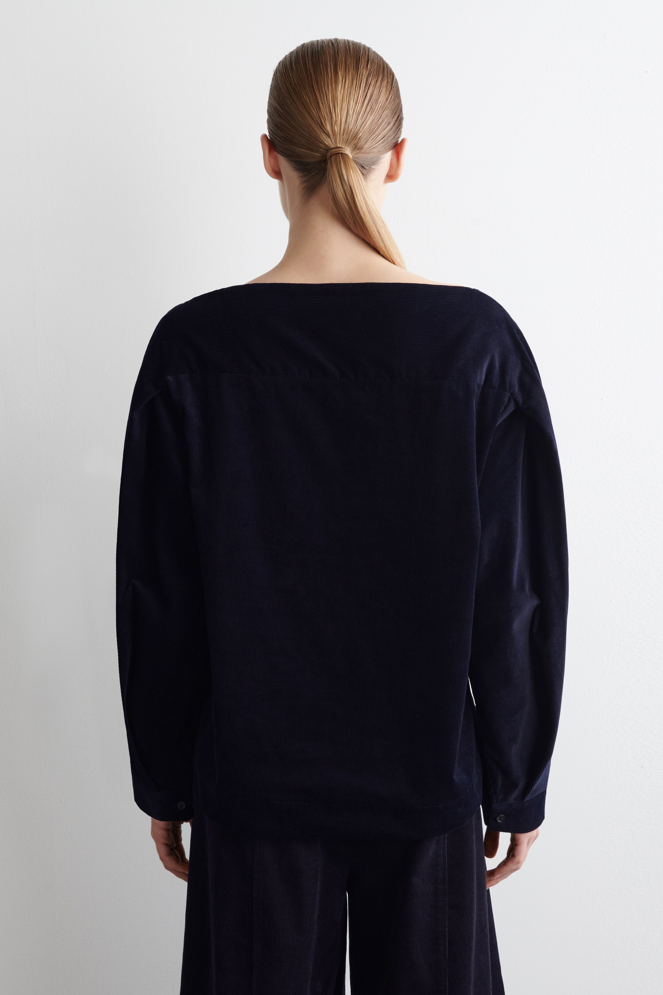 Visualizza immagine più grande: Un top in velluto a coste blu scuro viene presentato dal retro, caratterizzato da un'ampia scollatura a barca e maniche lunghe con polsini abbottonati. Il top è appoggiato su pantaloni a gamba larga in velluto a coste blu scuro coordinati, formando un insieme coordinato.