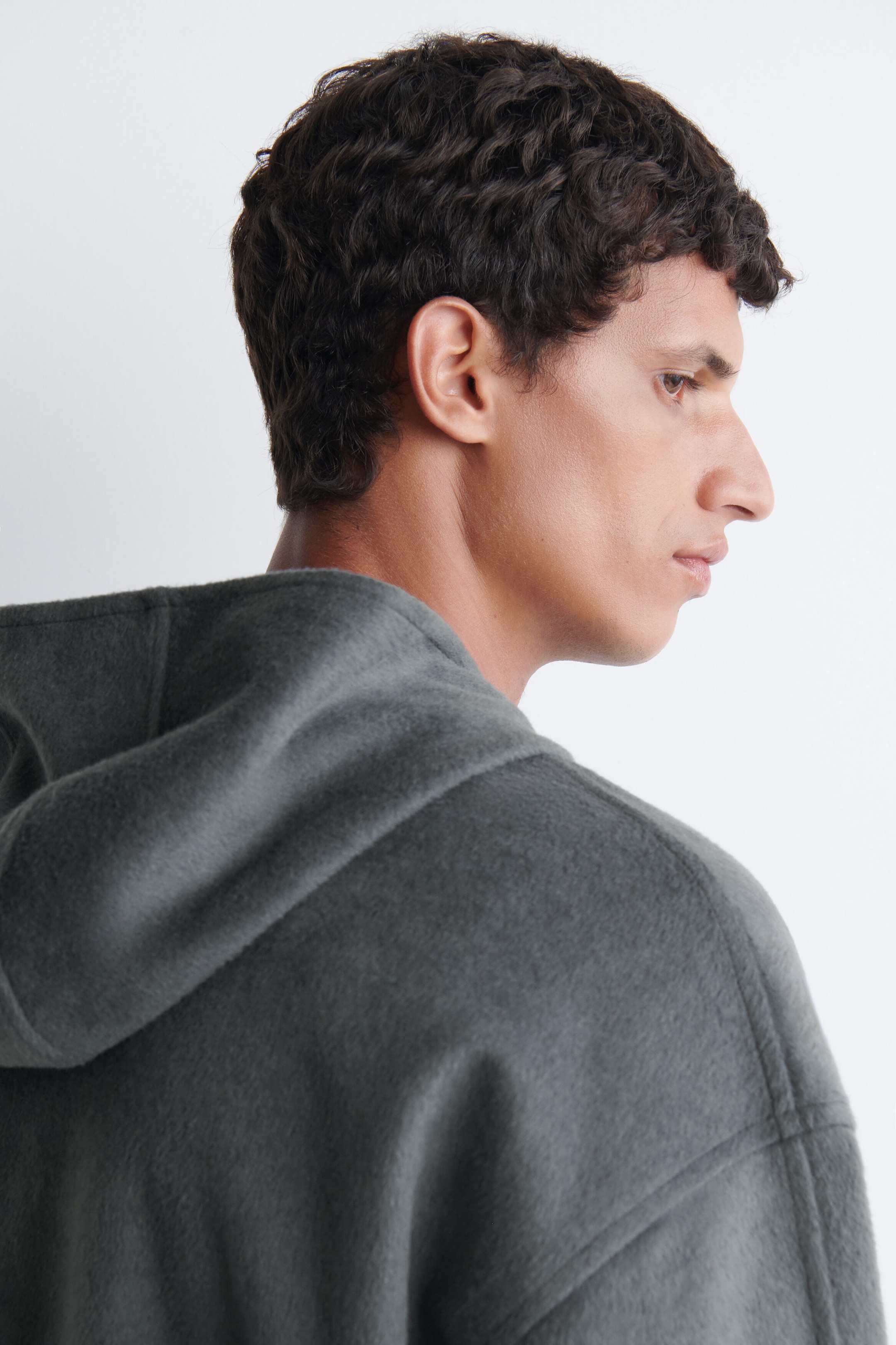 Un vêtement à capuchon gris foncé texturé est porté par le mannequin, présenté de profil. L’article est confectionné dans un tissu doux et brossé, avec une capuche reposant sur le dos. Un vêtement à capuchon gris foncé texturé est porté par le mannequin, présenté de profil. L’article est confectionné dans un tissu doux et brossé, avec une capuche reposant sur le dos.