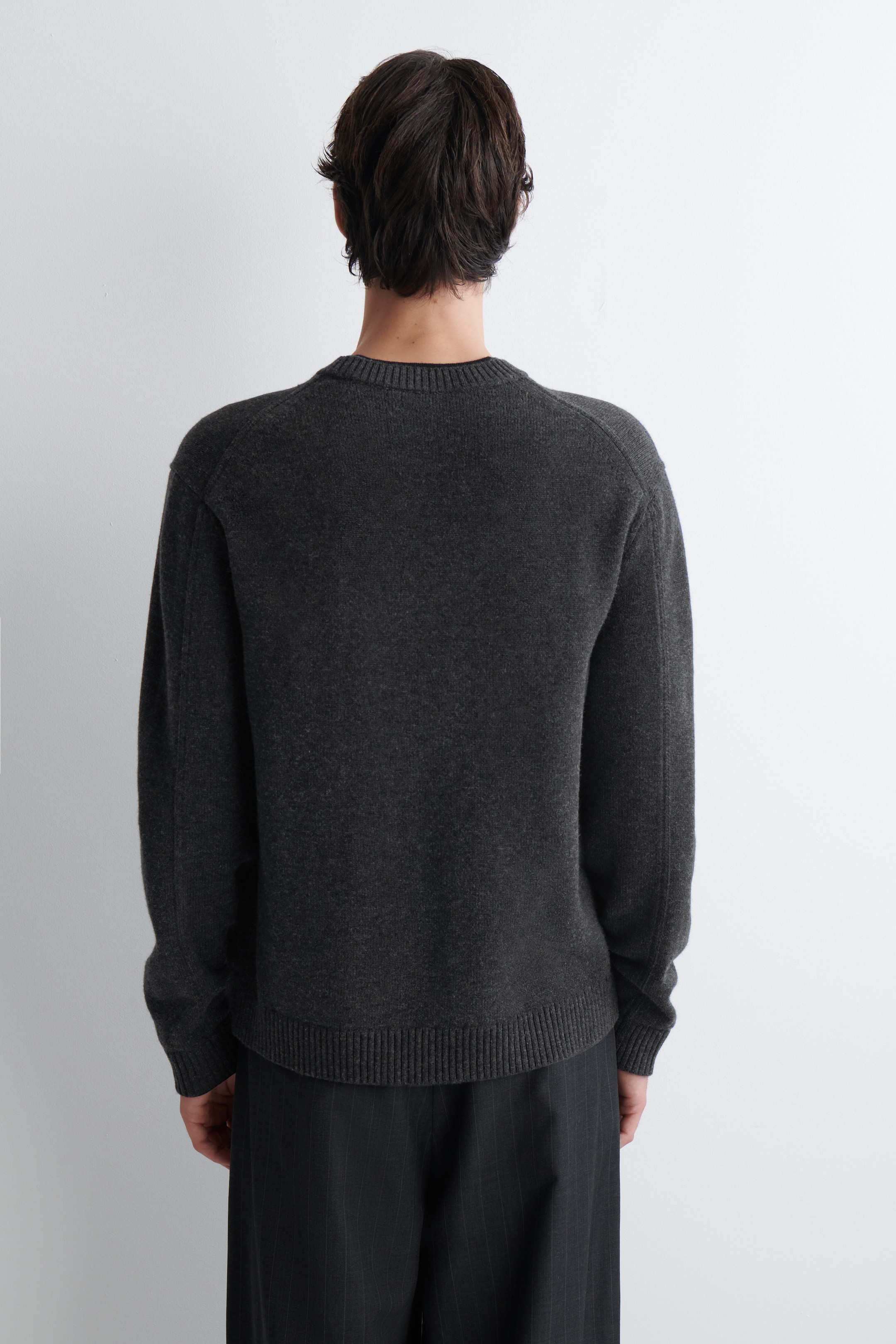 Grösseres Bild ansehen: Ein dunkelgrauer Pullover wird von hinten gezeigt, wobei sein lockerer Schnitt und die dezente Strickstruktur hervorgehoben werden. Er verfügt über einen gerippten Rundhalsausschnitt, lange Ärmel mit gerippten Bündchen und einen gerippten Saum.
