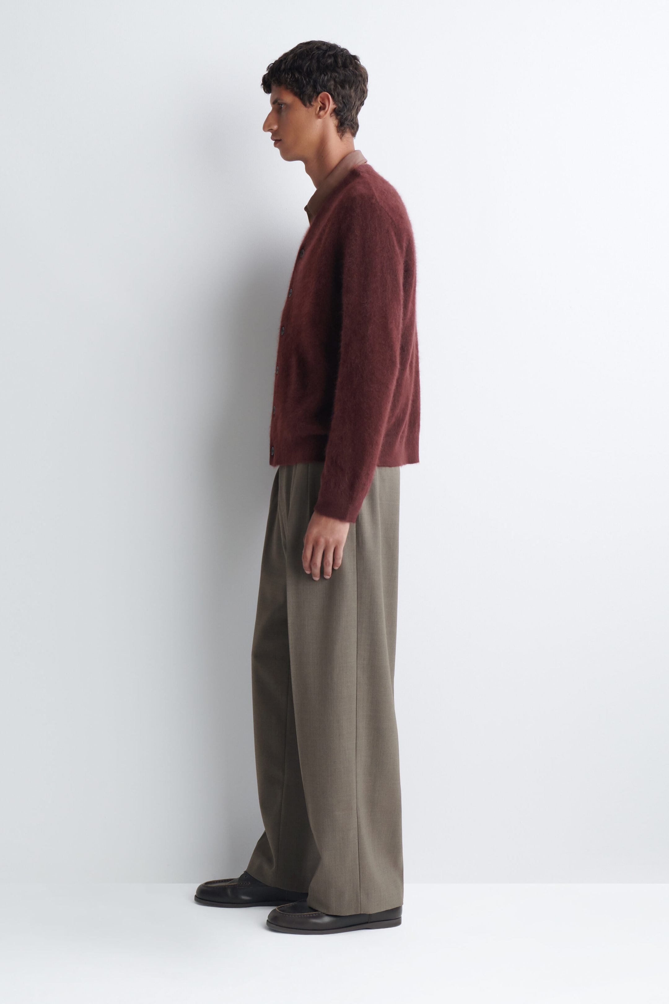 A brushed cashmere V-neck cardigan in a deep burgundy shade is worn by the model, who is standing in profile. The fuzzy-textured cardigan is buttoned up and layered over a brown collared shirt, paired with wide-leg taupe trousers. A brushed cashmere V-neck cardigan in a deep burgundy shade is worn by the model, who is standing in profile. The fuzzy-textured cardigan is buttoned up and layered over a brown collared shirt, paired with wide-leg taupe trousers.