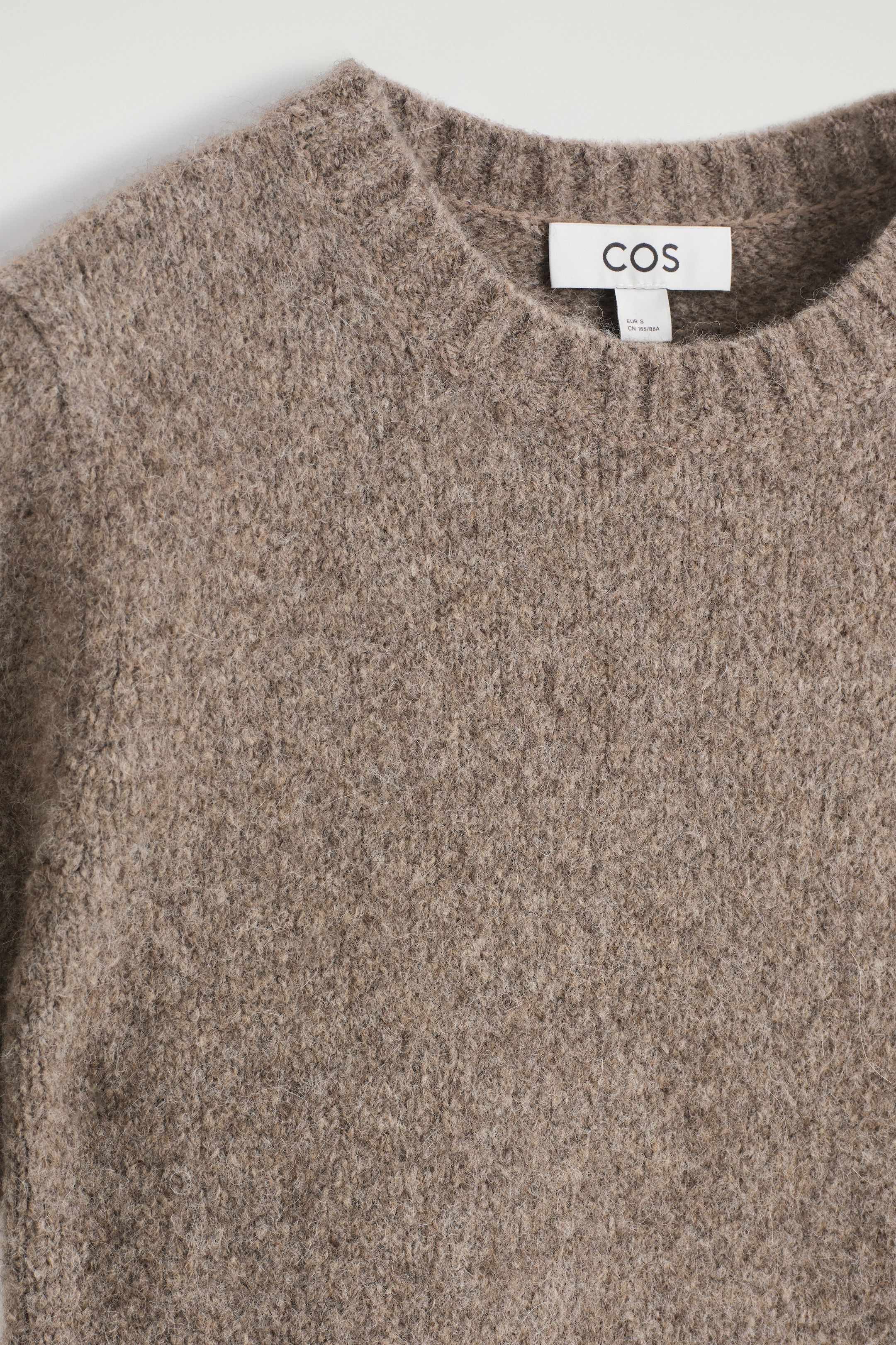 Agrandir l'image: Un pull chiné brun-gris est orienté vers l'avant, affichant une texture tricotée duveteuse et un col rond côtelé. Une étiquette de marque COS blanche est visible à l'arrière du cou.