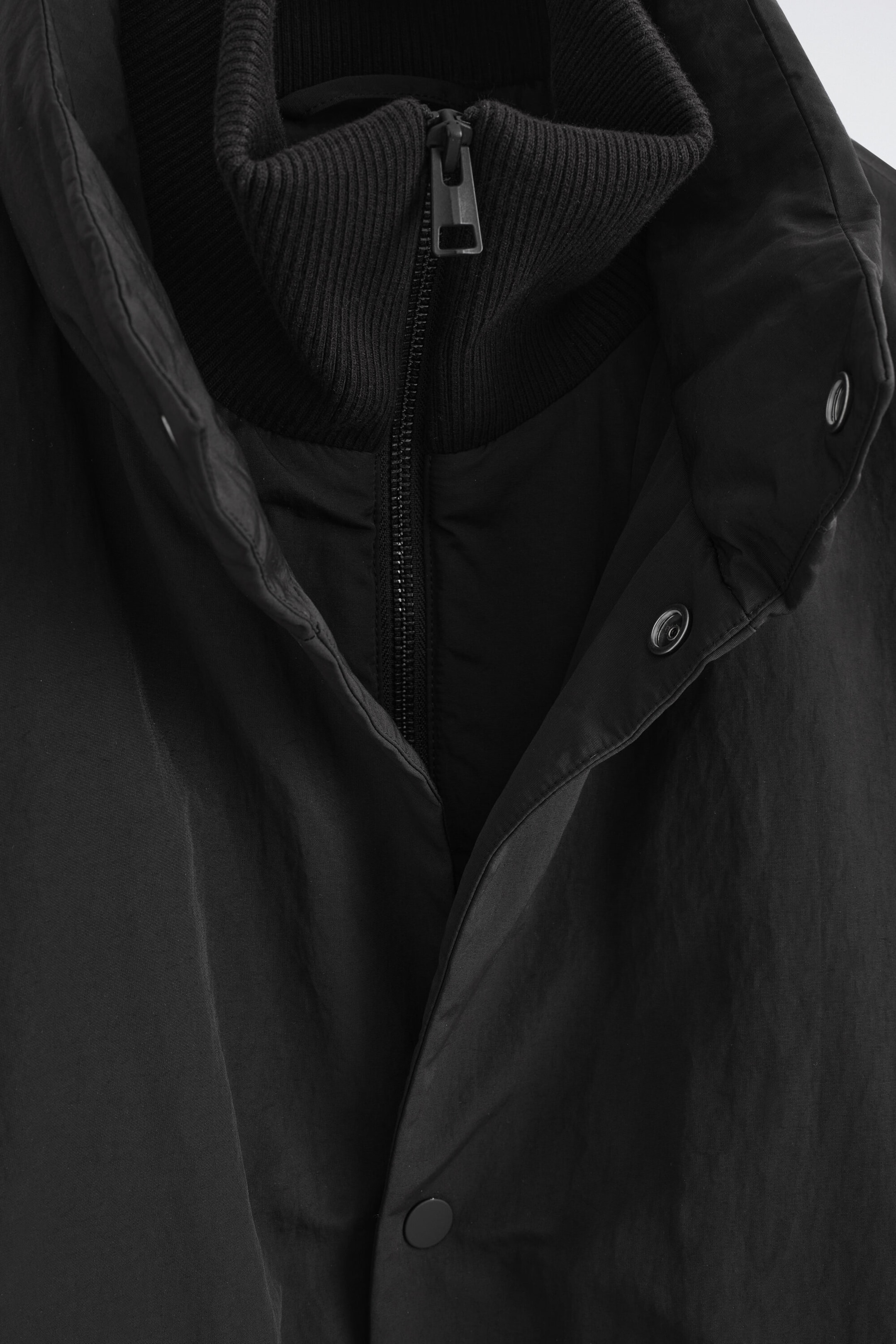 A close-up front view of a black padded funnel-neck down jacket highlighting its layered collar design. The jacket features a ribbed-knit inner collar with a metal zipper closure and an outer high-neck placket with snap-button fasteners. A close-up front view of a black padded funnel-neck down jacket highlighting its layered collar design. The jacket features a ribbed-knit inner collar with a metal zipper closure and an outer high-neck placket with snap-button fasteners.