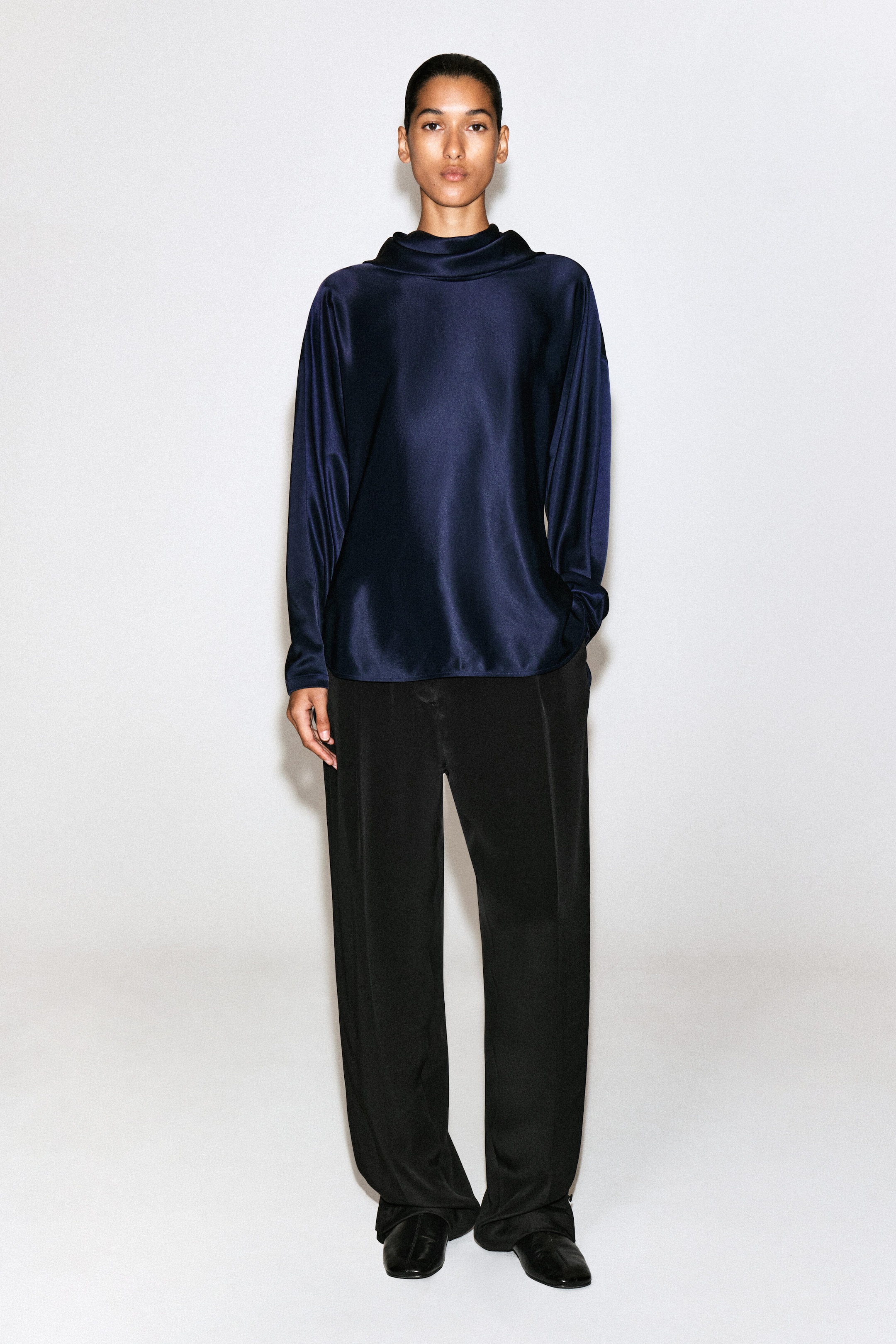 A dark navy cowl neck satin blouse is shown in a relaxed fit with a soft sheen in this front-facing view. The blouse features long sleeves and a draped, high cowl neck, falling loosely over black trousers.