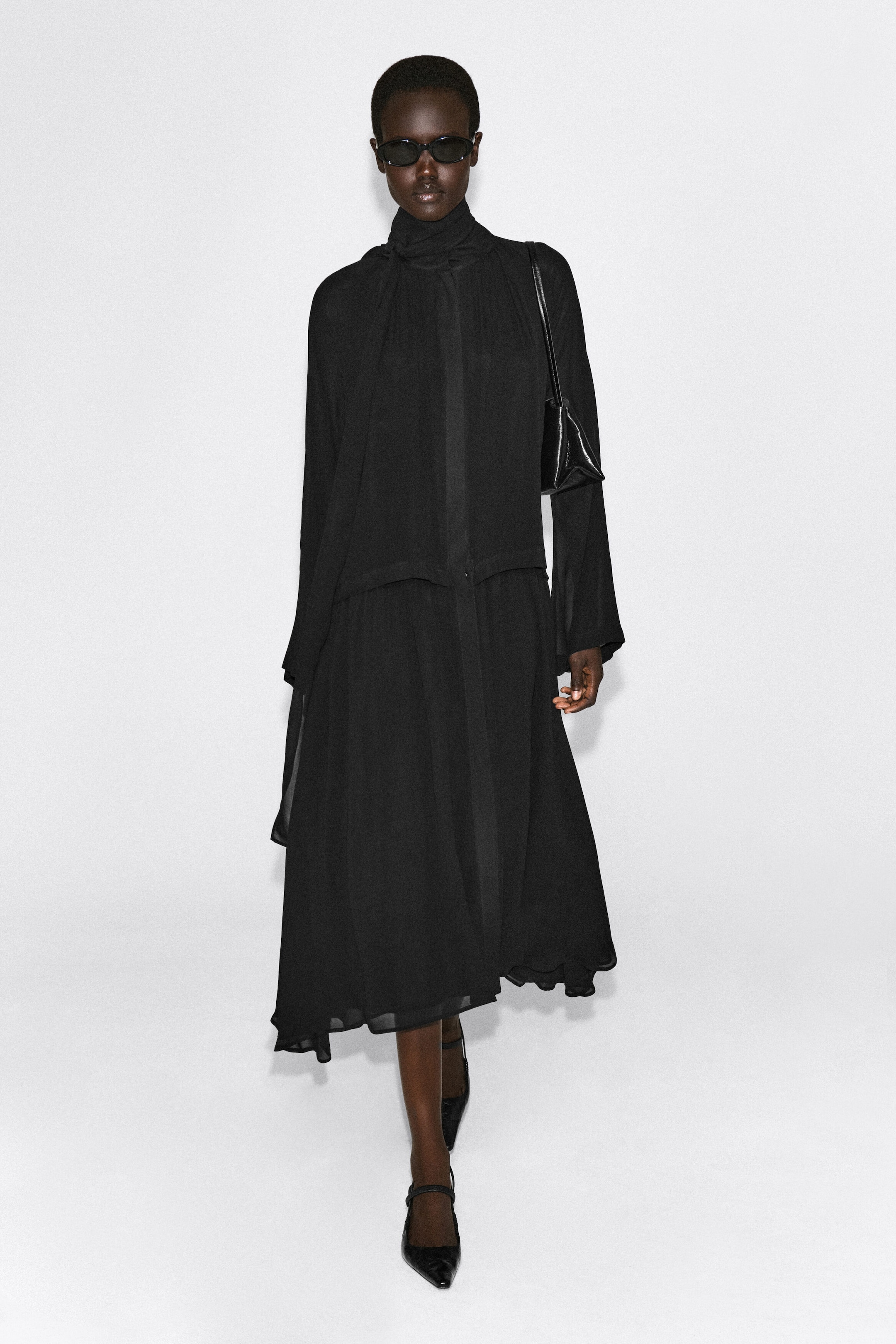 A black fluid midi dress with a scarf detail is shown from the front, featuring a high neckline with an integrated draped scarf and long, relaxed-fit sleeves. The dress flows into a layered, relaxed silhouette with a fluid asymmetrical midi-length hem.