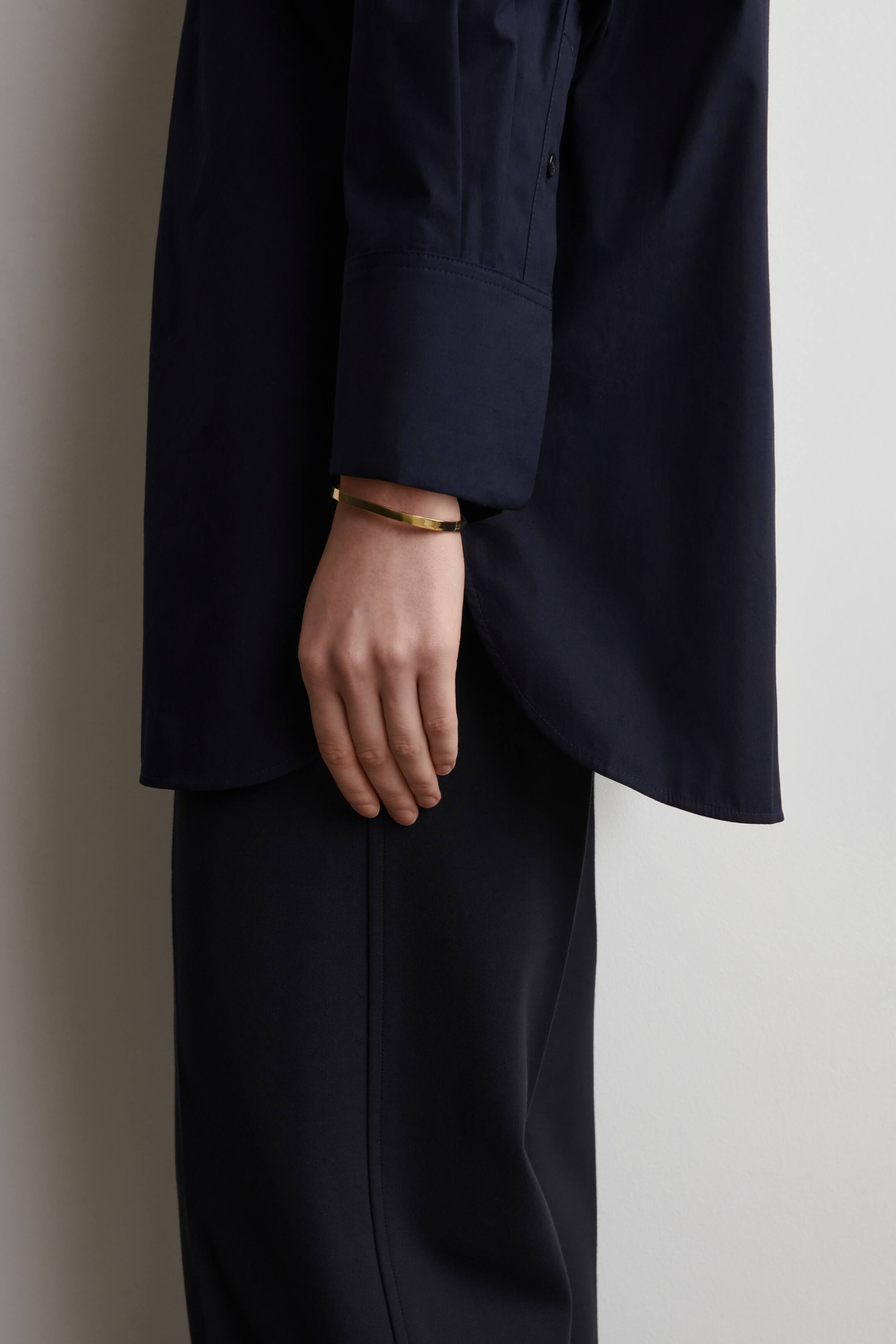 הצגת תמונה גדולה יותר: חולצה כחולה כהה עם שרוולים ארוכים, הכוללת חפתים רחבים וקו מכפלת מעוגל, נלבשת. צמיד דק בצבע זהב, פתוח בקצוות, נלבש על פרק כף היד מעל חפת החולצה, ומכנסיים כחולים כהים עם תפר קדמי אנכי נראים גם הם.