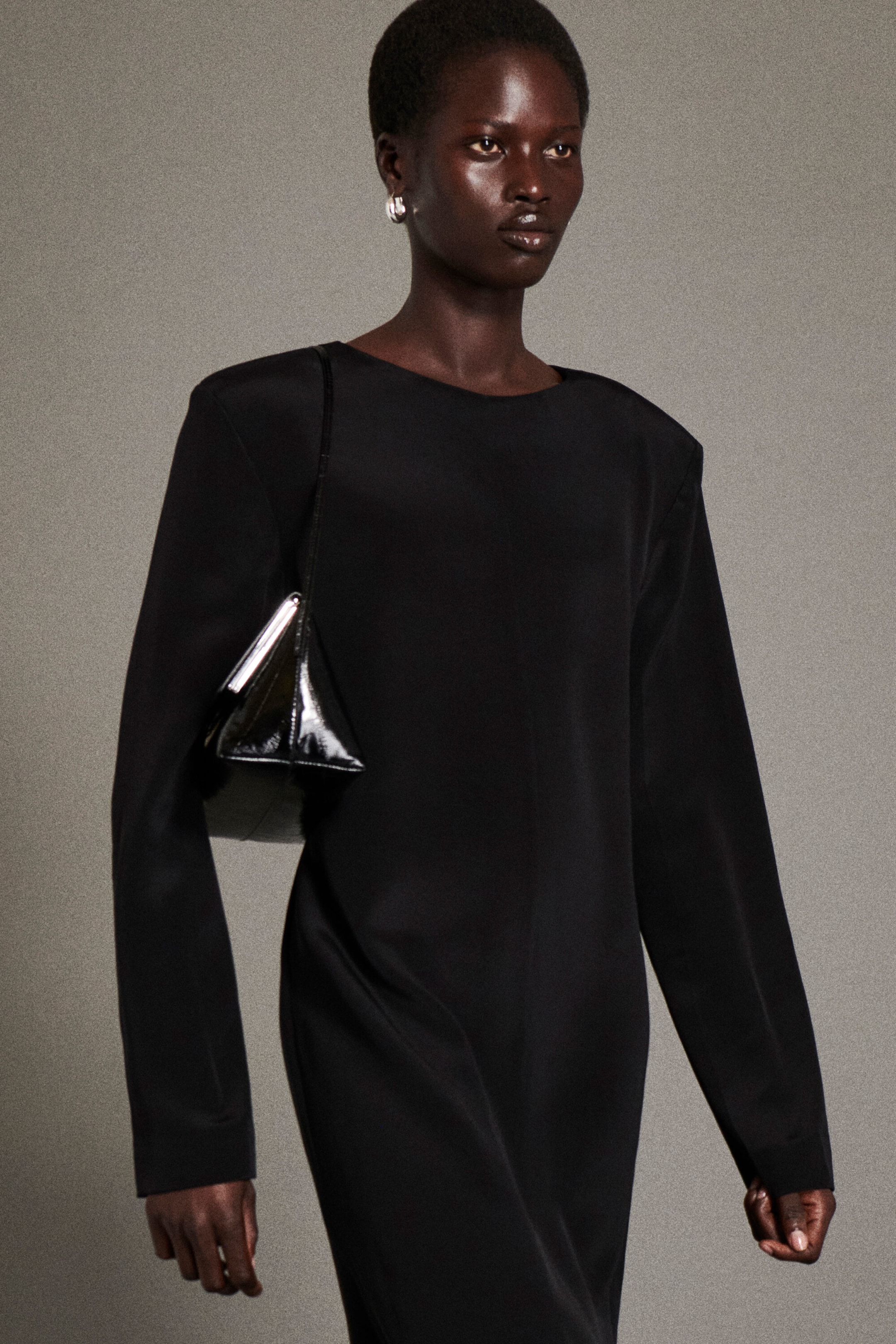 The button-detail silk crepe maxi dress is shown from the front, featuring a structured silhouette with padded shoulders, a round neckline, and long sleeves. The dress is styled with a black patent leather triangular bag worn over the shoulder. The button-detail silk crepe maxi dress is shown from the front, featuring a structured silhouette with padded shoulders, a round neckline, and long sleeves. The dress is styled with a black patent leather triangular bag worn over the shoulder.