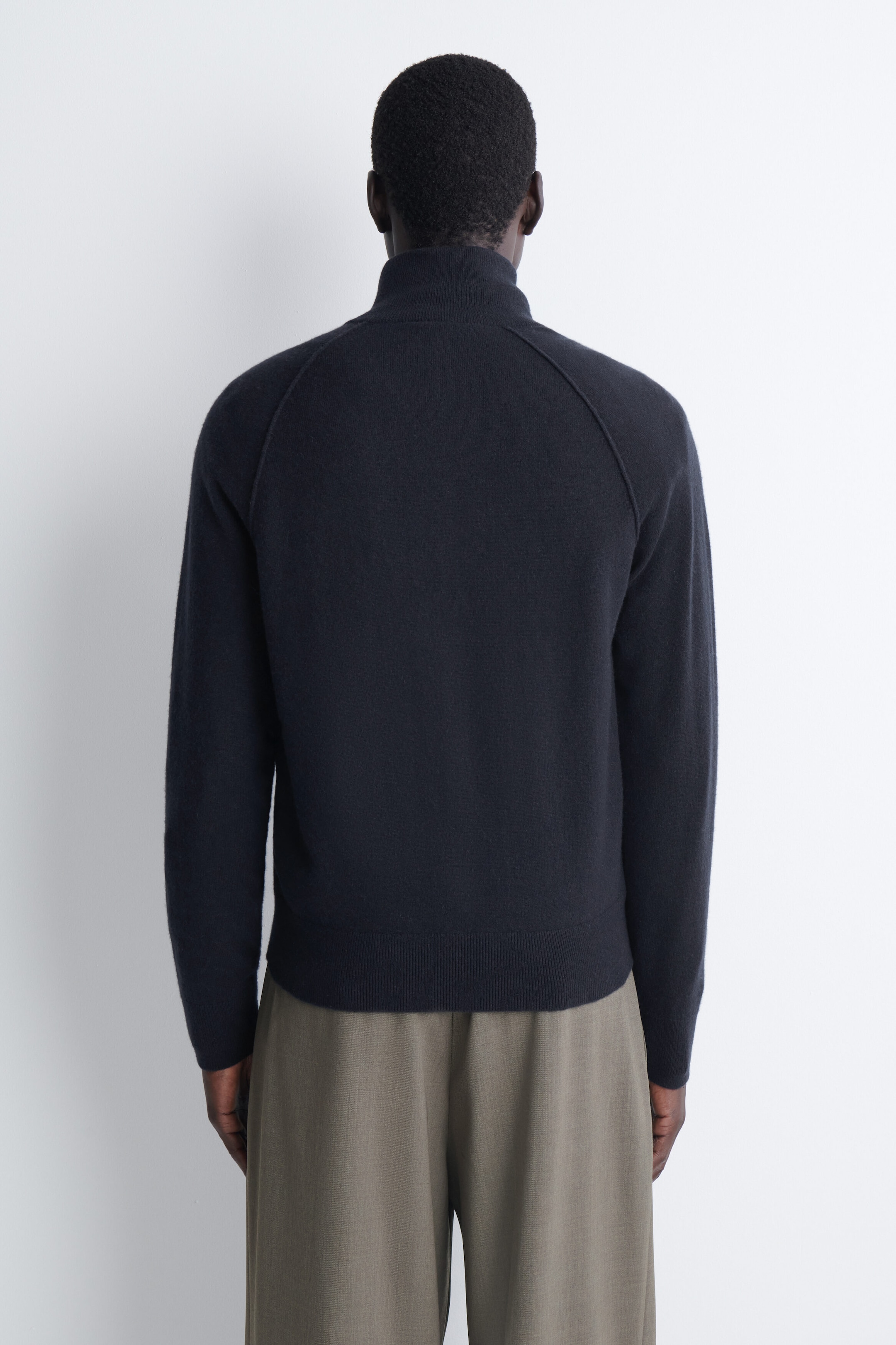 The cashmere funnel-neck zip-up cardigan is shown from the back, highlighting its dark navy knit construction, raglan sleeves, and a high funnel neck. The cardigan features a ribbed hem and is paired with loose-fitting trousers to demonstrate its relaxed yet structured fit. The cashmere funnel-neck zip-up cardigan is shown from the back, highlighting its dark navy knit construction, raglan sleeves, and a high funnel neck. The cardigan features a ribbed hem and is paired with loose-fitting trousers to demonstrate its relaxed yet structured fit.