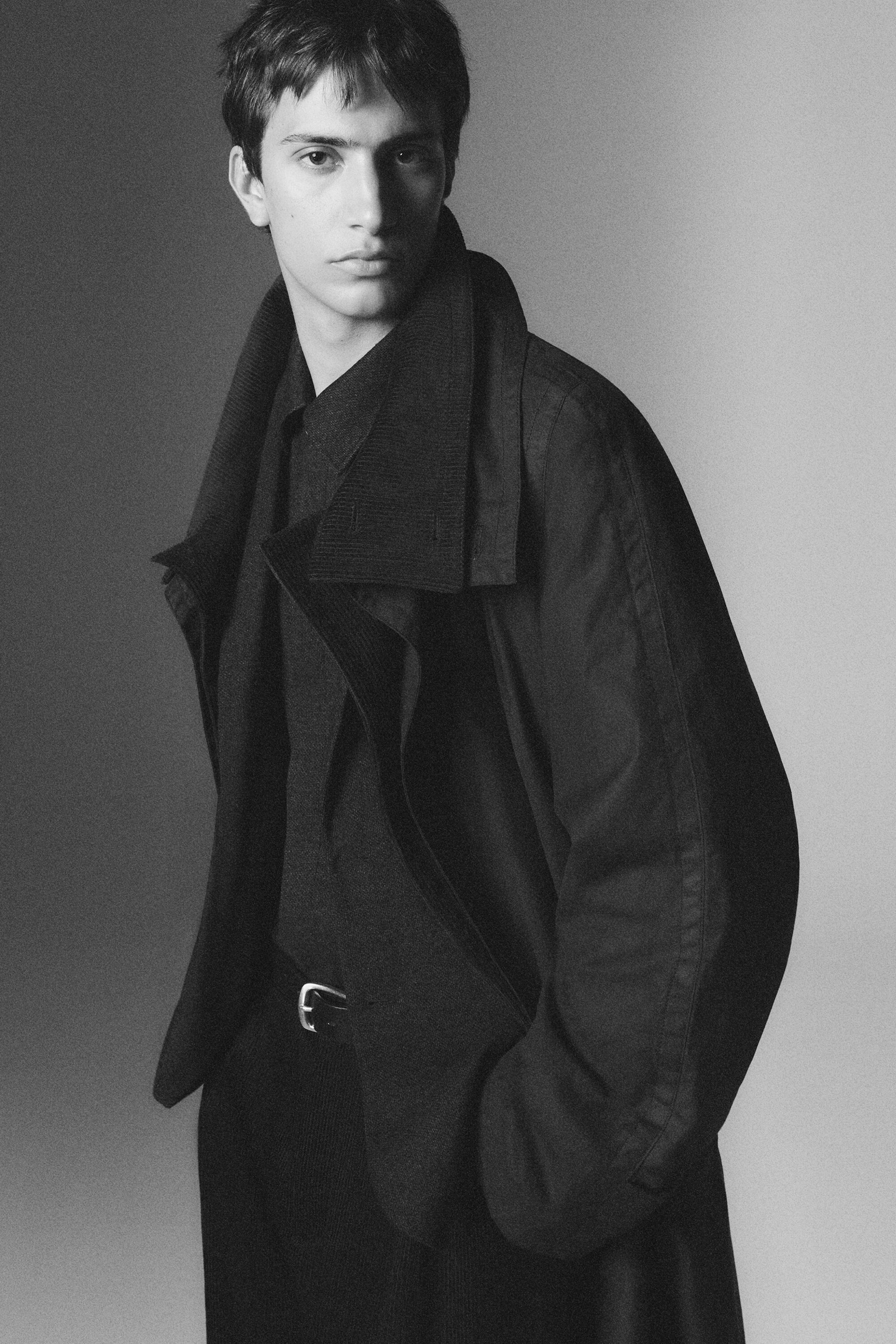The dark waxed-cotton trench coat features a relaxed silhouette with a wide, textured corduroy collar. It is shown in a three-quarter front view, worn open over a dark collared shirt and trousers with a silver belt buckle. The dark waxed-cotton trench coat features a relaxed silhouette with a wide, textured corduroy collar. It is shown in a three-quarter front view, worn open over a dark collared shirt and trousers with a silver belt buckle.