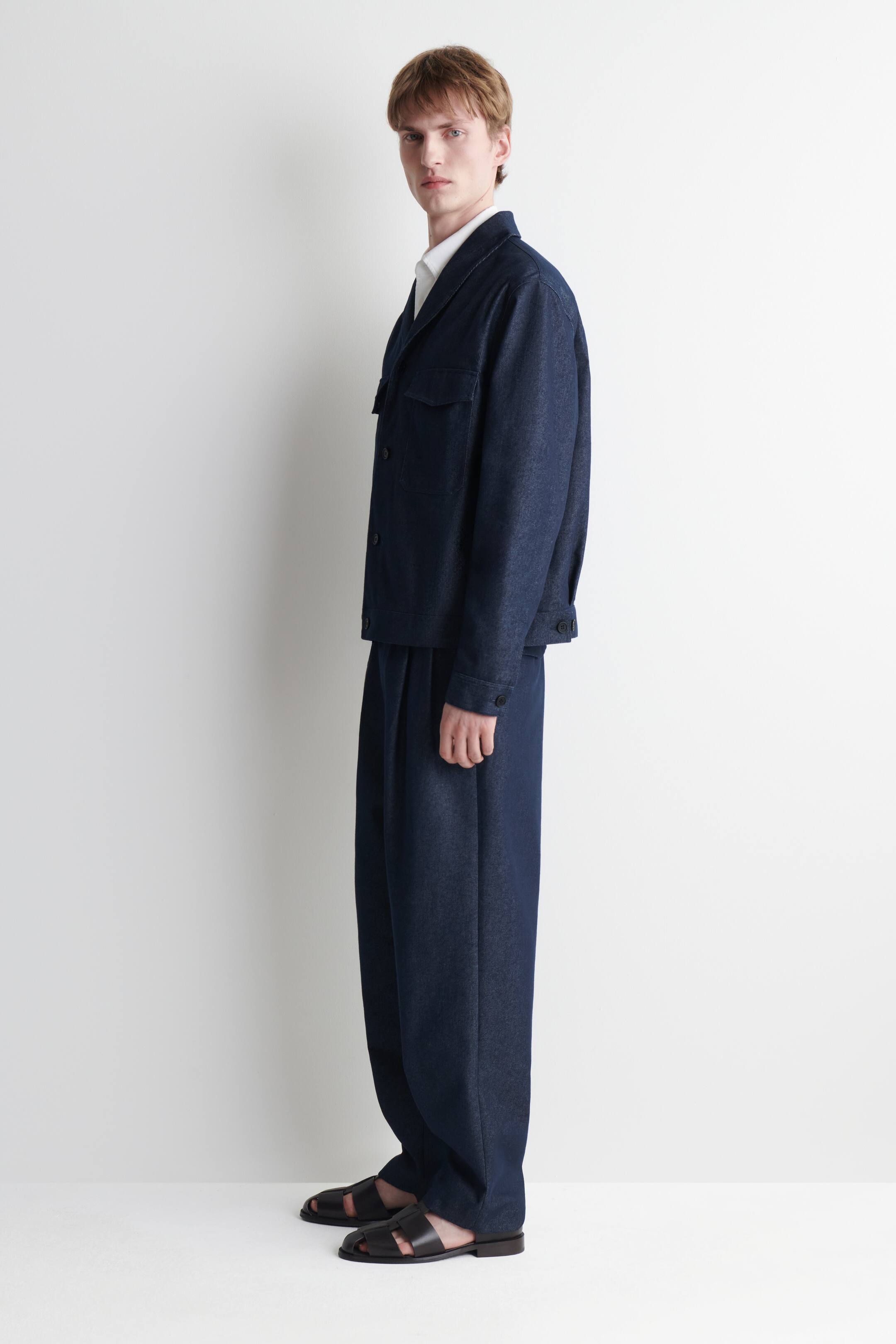 Visualizza immagine più grande: Una giacca in denim blu scuro e i pantaloni coordinati a gamba larga vengono presentati in una vista di tre quarti, con la giacca caratterizzata da un colletto a punta, due tasche con patta sul petto e polsini con bottoni sopra una camicia bianca con colletto. I pantaloni dalla vestibilità morbida mostrano pieghe visibili, mentre sandali neri in pelle con struttura a gabbia completano l'ensemble.