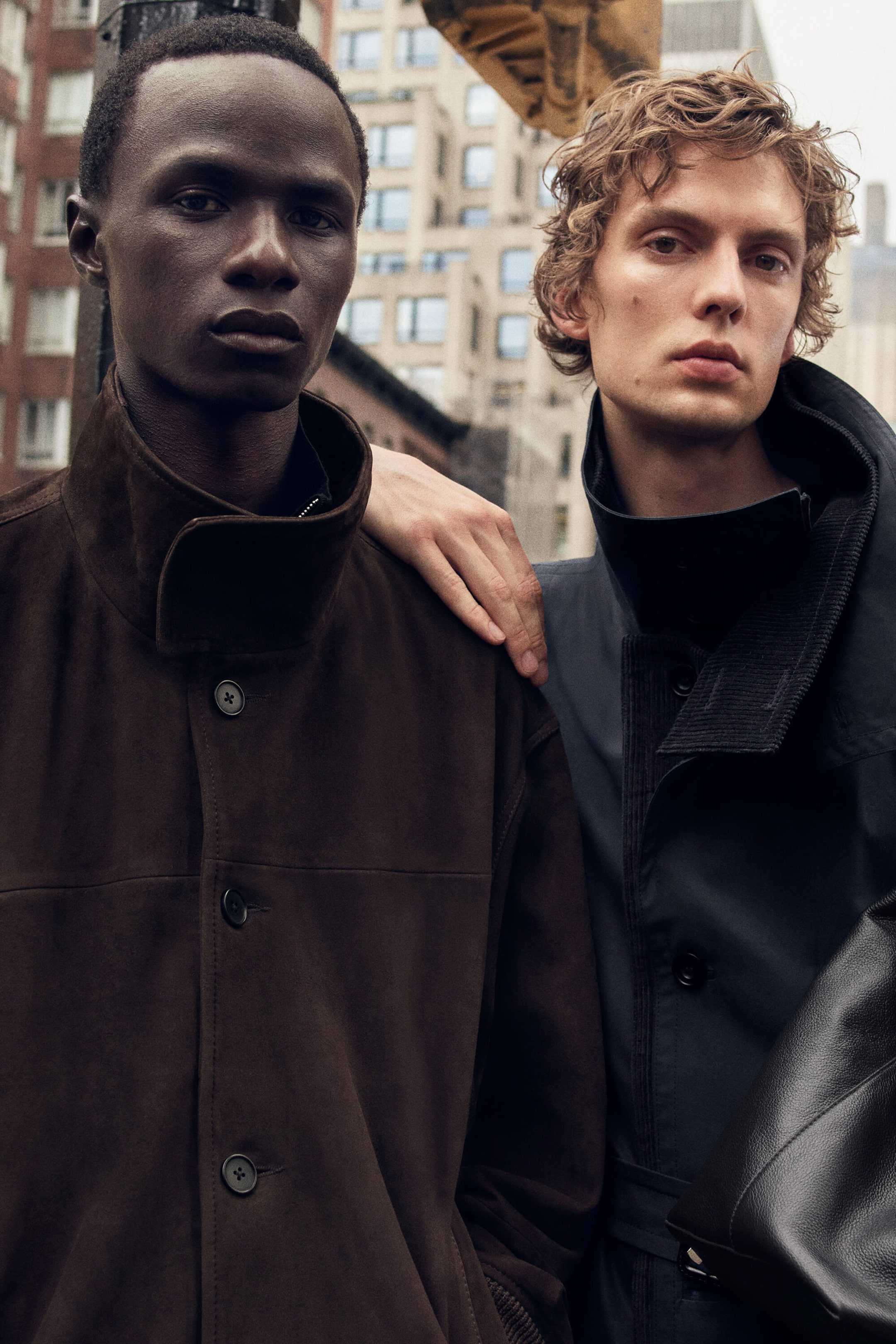 Zwei Herren-Oberbekleidungsstücke sind von vorne dargestellt: links eine dunkelbraune Wildlederjacke und rechts ein marineblauer Technical Coat. Die braune Wildlederjacke ist mit einem hohen Stehkragen und einer Knopfleiste vorne gestaltet, während der marineblaue Mantel einen mehrlagigen Kragen mit strukturierter Innendetailierung aufweist.
