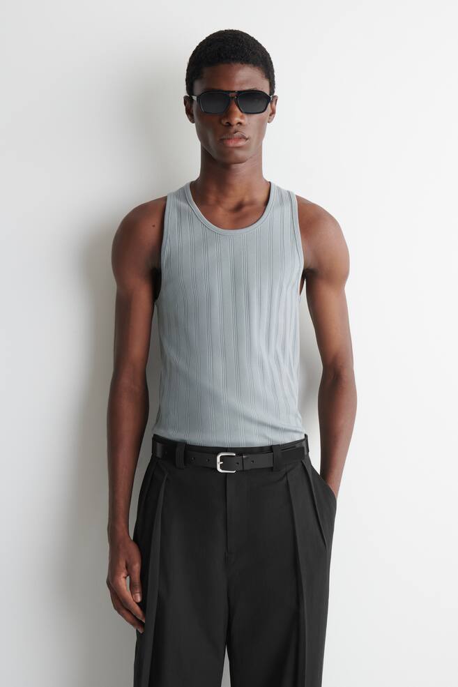 Ein hellgraues, geripptes Tanktop wird von einem Model getragen und zeigt seine schmale Passform und den Rundhalsausschnitt. Das Tanktop ist in eine schwarze Hose gesteckt, die mit einem schwarzen GÃ¼rtel gesichert ist. - 1