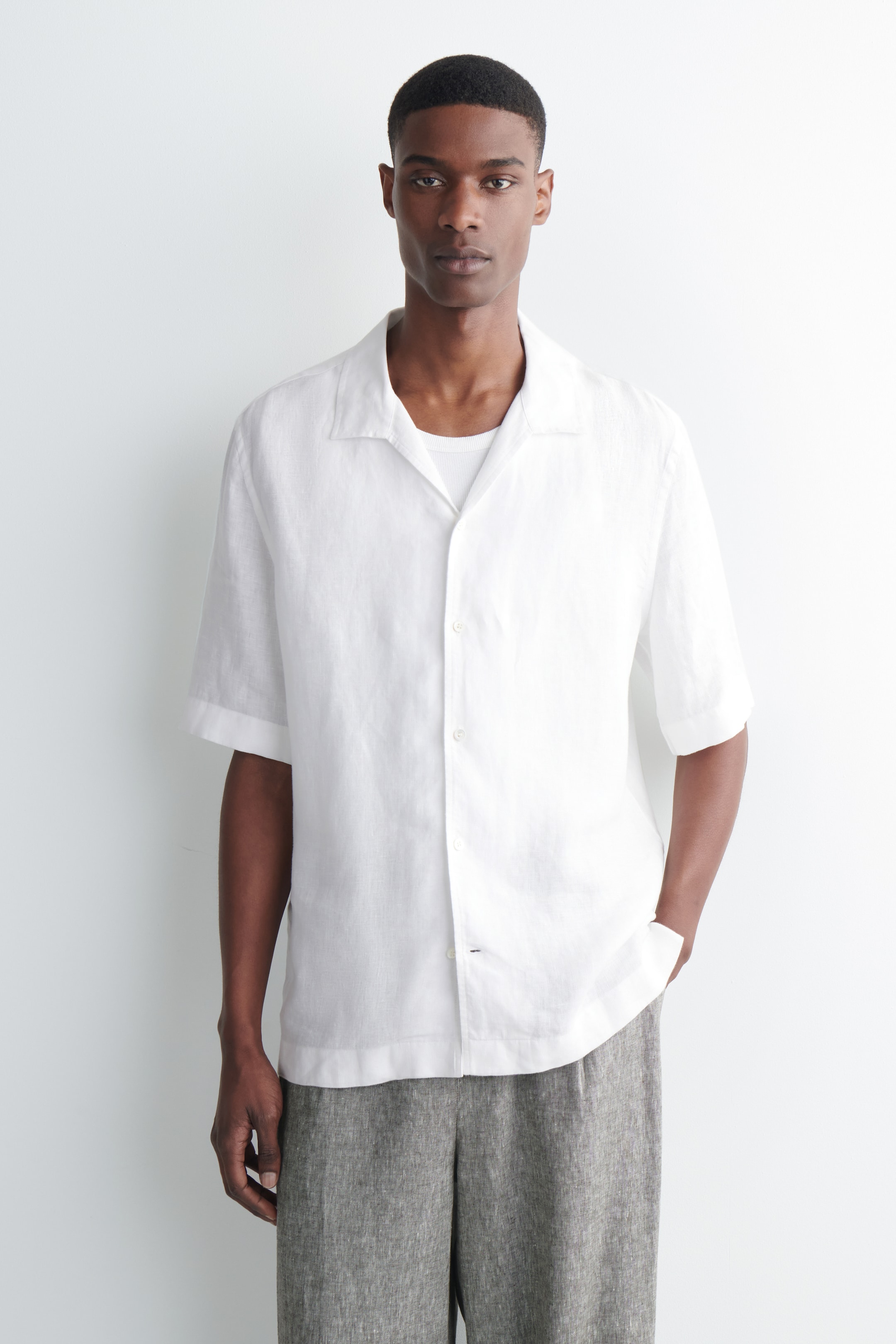 View larger image: A white linen short-sleeved shirt, featuring a Cuban collar and visible white buttons down the front, is styled open over a white ribbed vest.