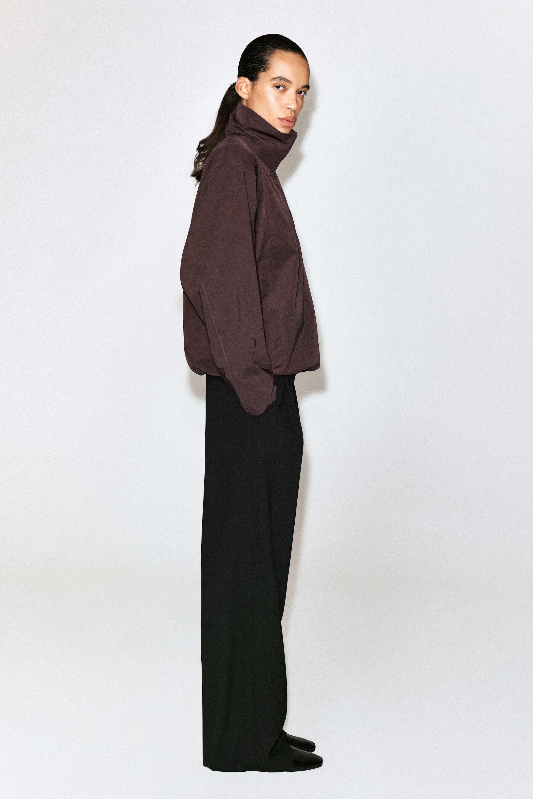 This dark brown high-neck jacket is shown facing right, highlighting its structured profile, high funnel collar, and gathered drawstring hem that creates a rounded silhouette. It is paired with black wide-leg trousers and black shoes. This dark brown high-neck jacket is shown facing right, highlighting its structured profile, high funnel collar, and gathered drawstring hem that creates a rounded silhouette. It is paired with black wide-leg trousers and black shoes.