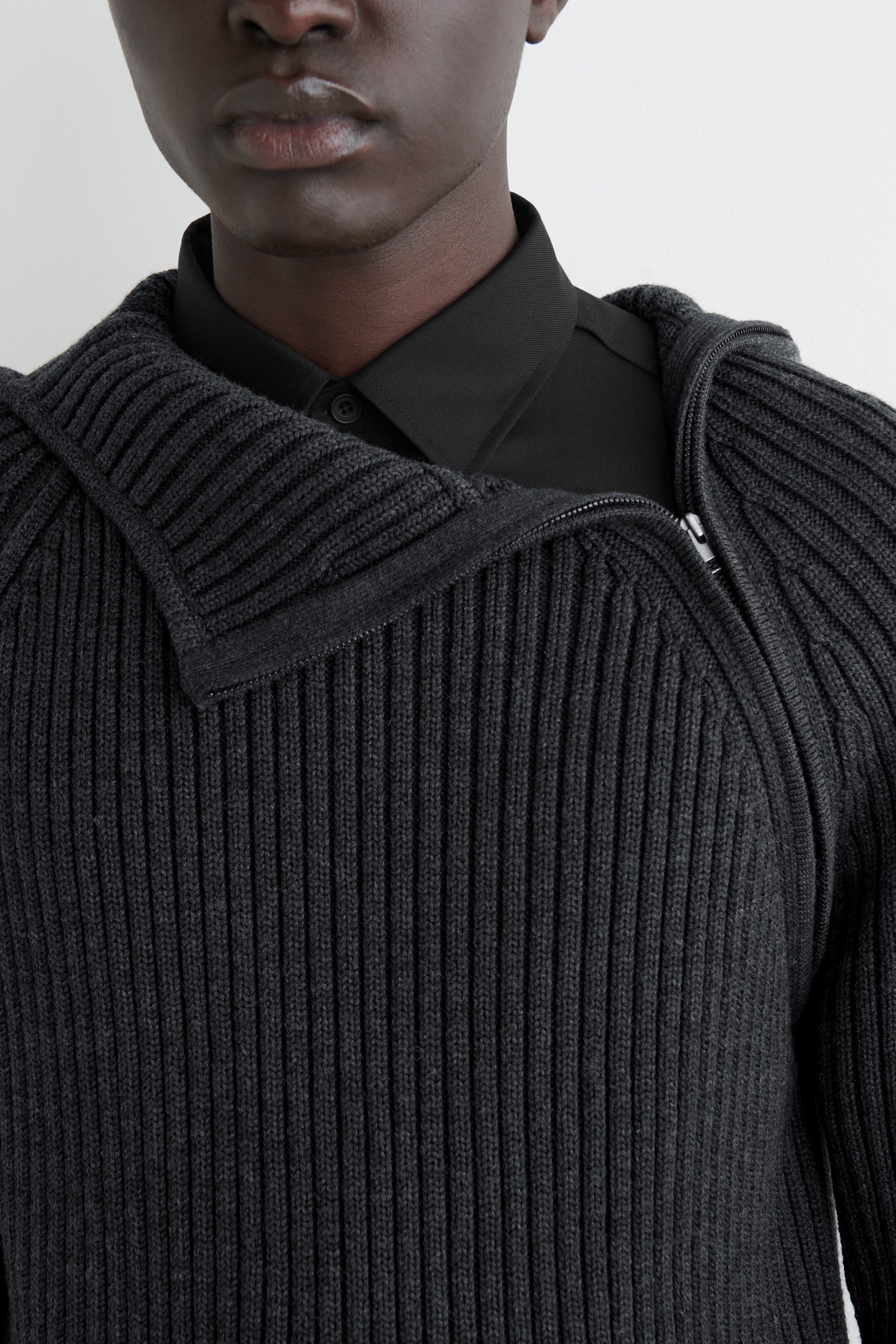 Agrandir l'image: Un vêtement en tricot côtelé gris foncé est présenté de face, avec un col large et plié, et une fermeture éclair diagonale argentée. Il est porté sur une chemise noire à col, mettant en valeur son encolure unique et sa texture.