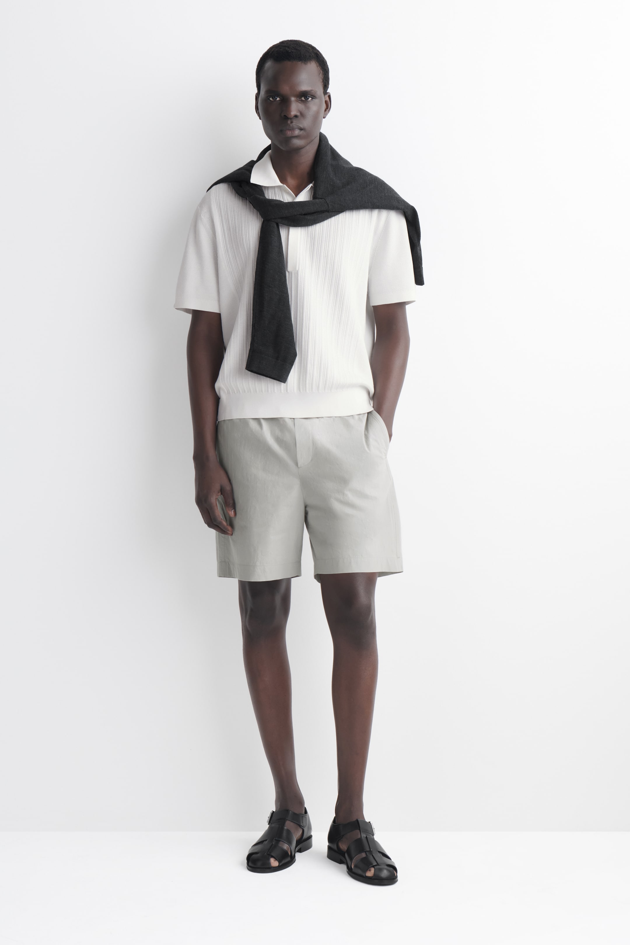 Ver imagen más grande: Se lleva puesta una camiseta polo de color crema claro, finamente acanalada, que presenta mangas cortas, un cuello clásico y un dobladillo acanalado. Un suéter oscuro está colocado sobre sus hombros.