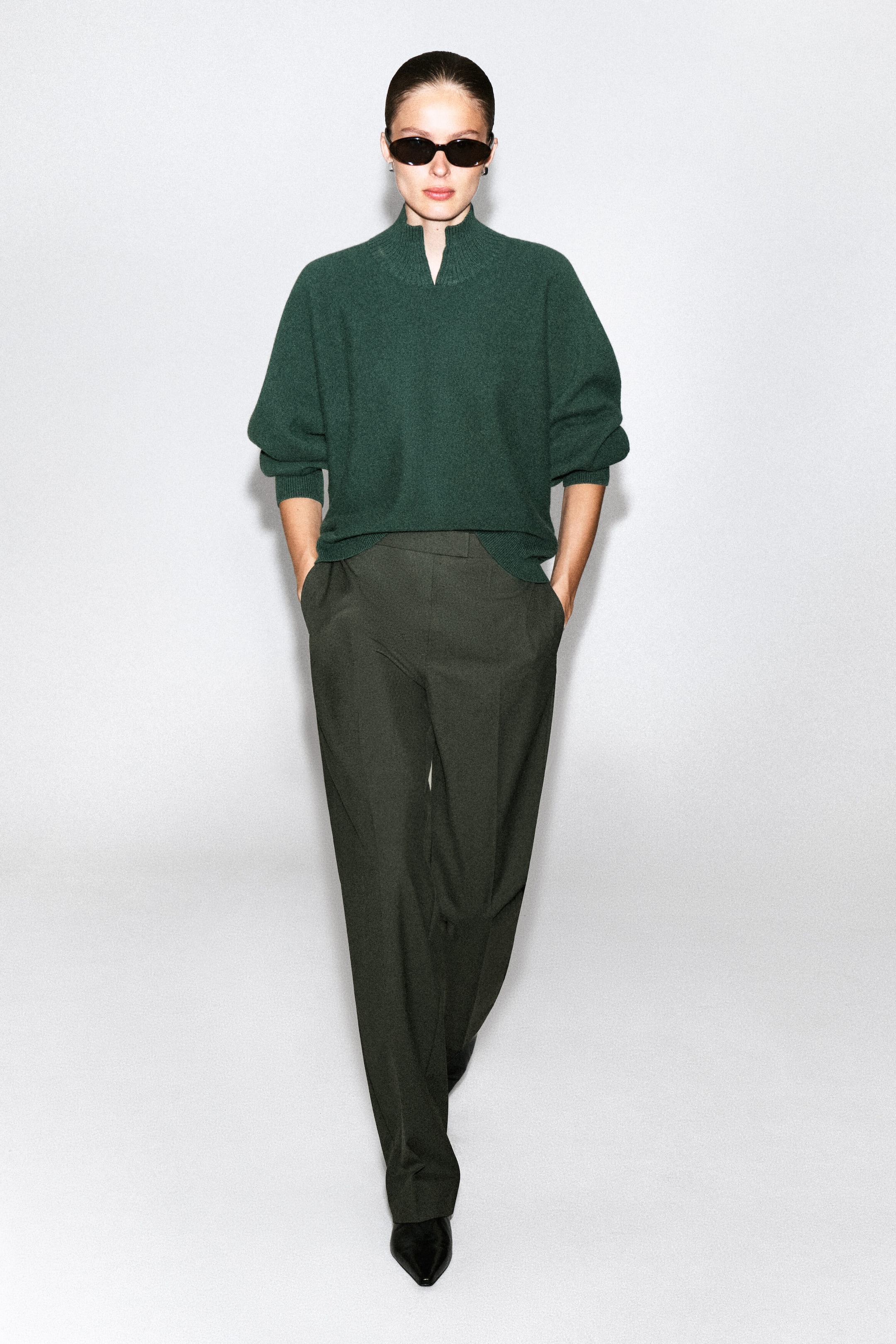 Un pull en laine bouillie vert foncé avec un col cheminée côtelé et une découpe en V sur le devant est porté par le modèle. Le chandail coupe décontractée est agencé avec un pantalon foncé et des lunettes de soleil noires. Un pull en laine bouillie vert foncé avec un col cheminée côtelé et une découpe en V sur le devant est porté par le modèle. Le chandail coupe décontractée est agencé avec un pantalon foncé et des lunettes de soleil noires.