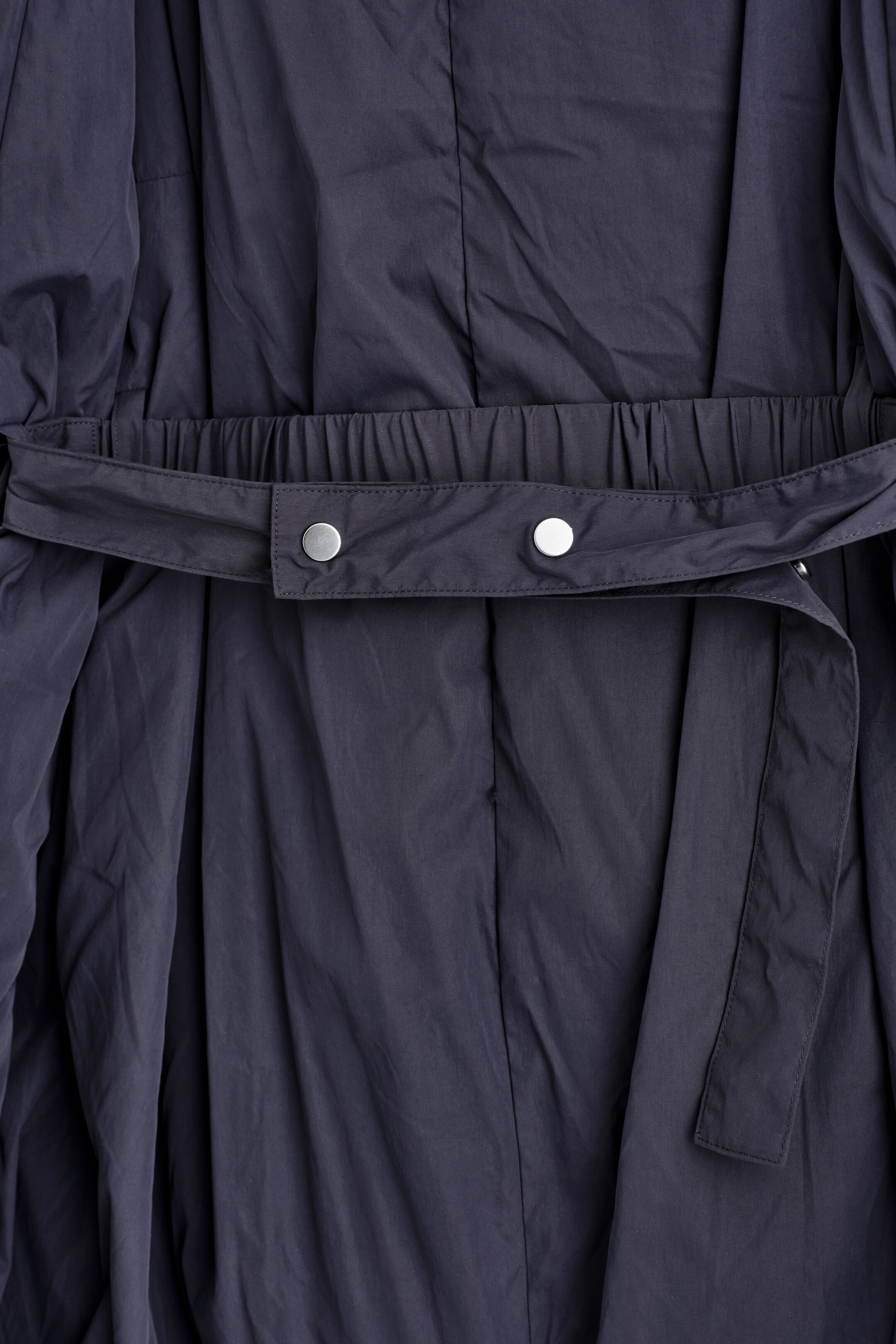 Ver imagen más grande: Una prenda de color gris oscuro presenta una cintura ceñida, con un cinturón de tela a juego que la envuelve. Este cinturón muestra dos broches a presión de tono plateado visibles en su sección FRONTAL, mientras que el material de la prenda se recoge y cae suavemente por encima y por debajo de la cintura.