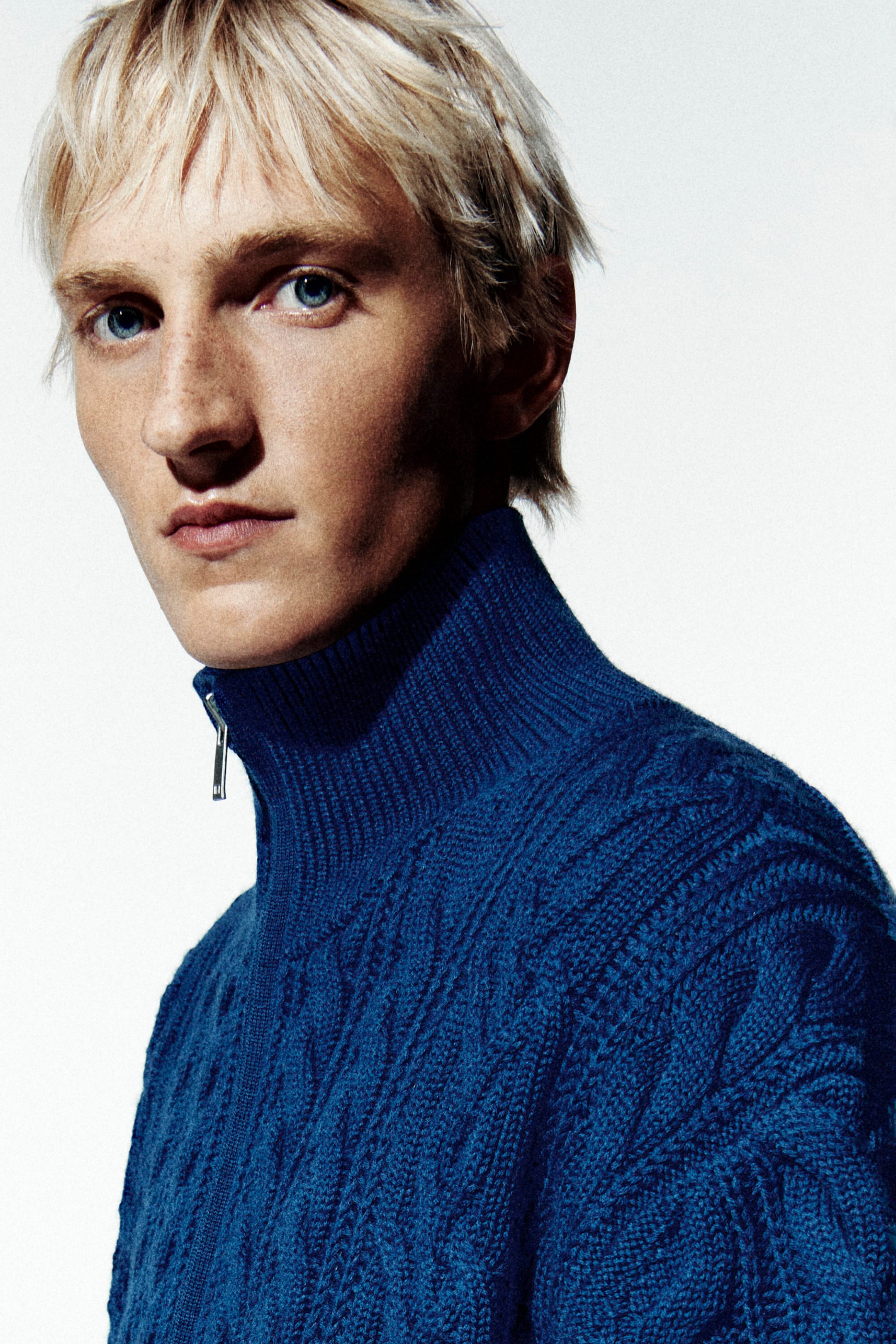 A blue cable-knit wool funnel-neck zip-up jacket is shown in a close-up, three-quarter front view, highlighting its high ribbed collar with a silver-tone zipper pull. The jacket features a thick, textured cable-knit pattern across the shoulders and body. A blue cable-knit wool funnel-neck zip-up jacket is shown in a close-up, three-quarter front view, highlighting its high ribbed collar with a silver-tone zipper pull. The jacket features a thick, textured cable-knit pattern across the shoulders and body.