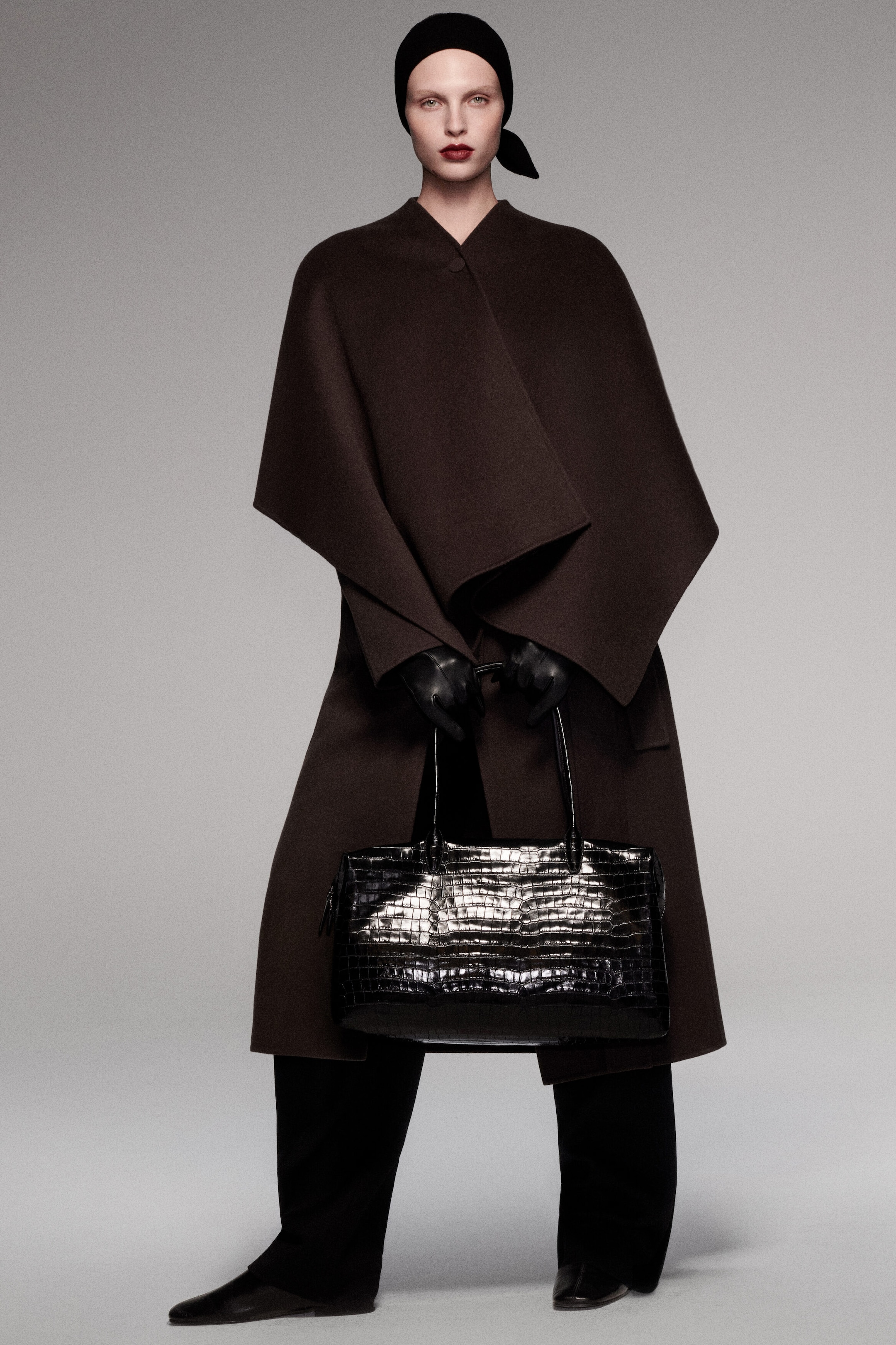 A model facing forward is wearing a dark brown double-faced wool-blend cape with structured, draped folds layered over a matching belted double-faced wool-blend long coat. They are holding a glossy black croc-effect leather Frieze bowling bag by its dual top handles in front. A model facing forward is wearing a dark brown double-faced wool-blend cape with structured, draped folds layered over a matching belted double-faced wool-blend long coat. They are holding a glossy black croc-effect leather Frieze bowling bag by its dual top handles in front.