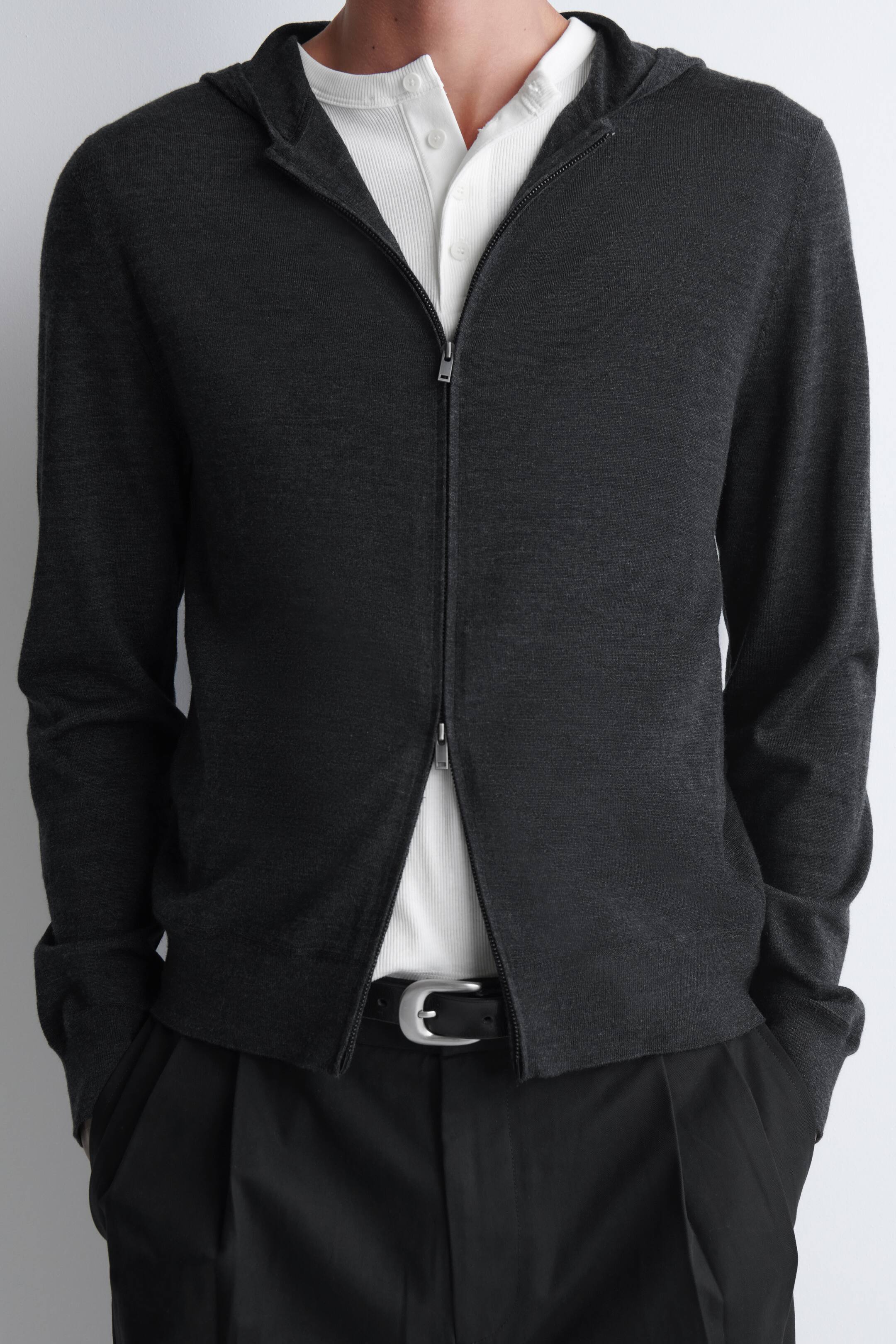 Agrandir l'image: Un sweat à capuche zippé en laine mérinos gris charbon est porté par le mannequin, avec une fermeture éclair argentée à double sens partiellement fermée au centre. Le sweat à capuche est superposé sur un henley côtelé blanc et associé à un pantalon noir.