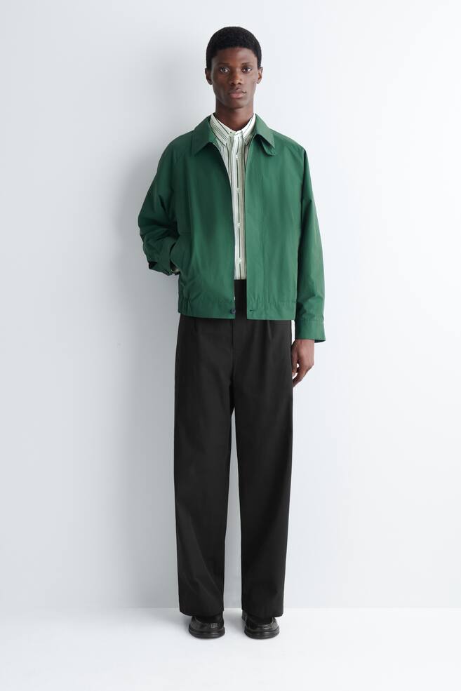 The model is wearing a green collared jacket with an elasticated hem and cuffs. One hand is placed in a jacket pocket, revealing a striped shirt underneath. - 1