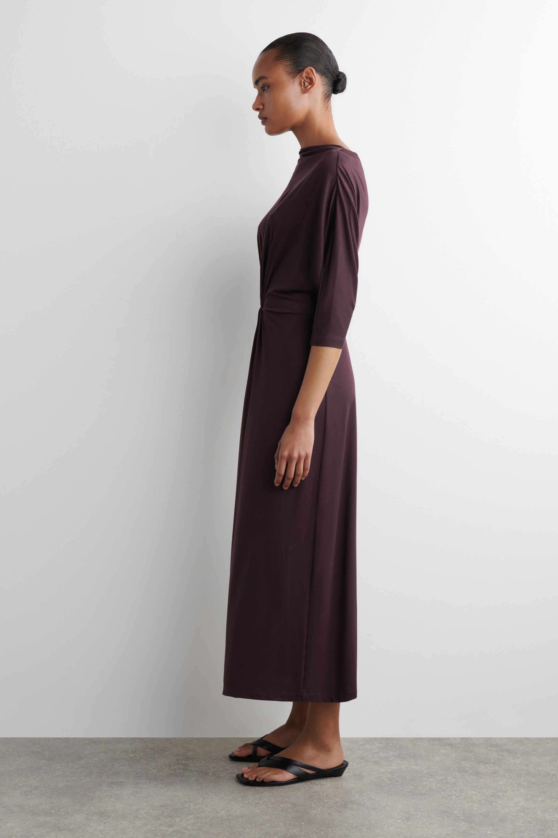 Agrandir l'image: Une robe maxi de couleur prune foncé est présentée de profil, avec un large col bateau et des manches trois-quarts. Un détail drapé et torsadé accentue le côté de la taille, avec la jupe tombant librement jusqu'aux chevilles.