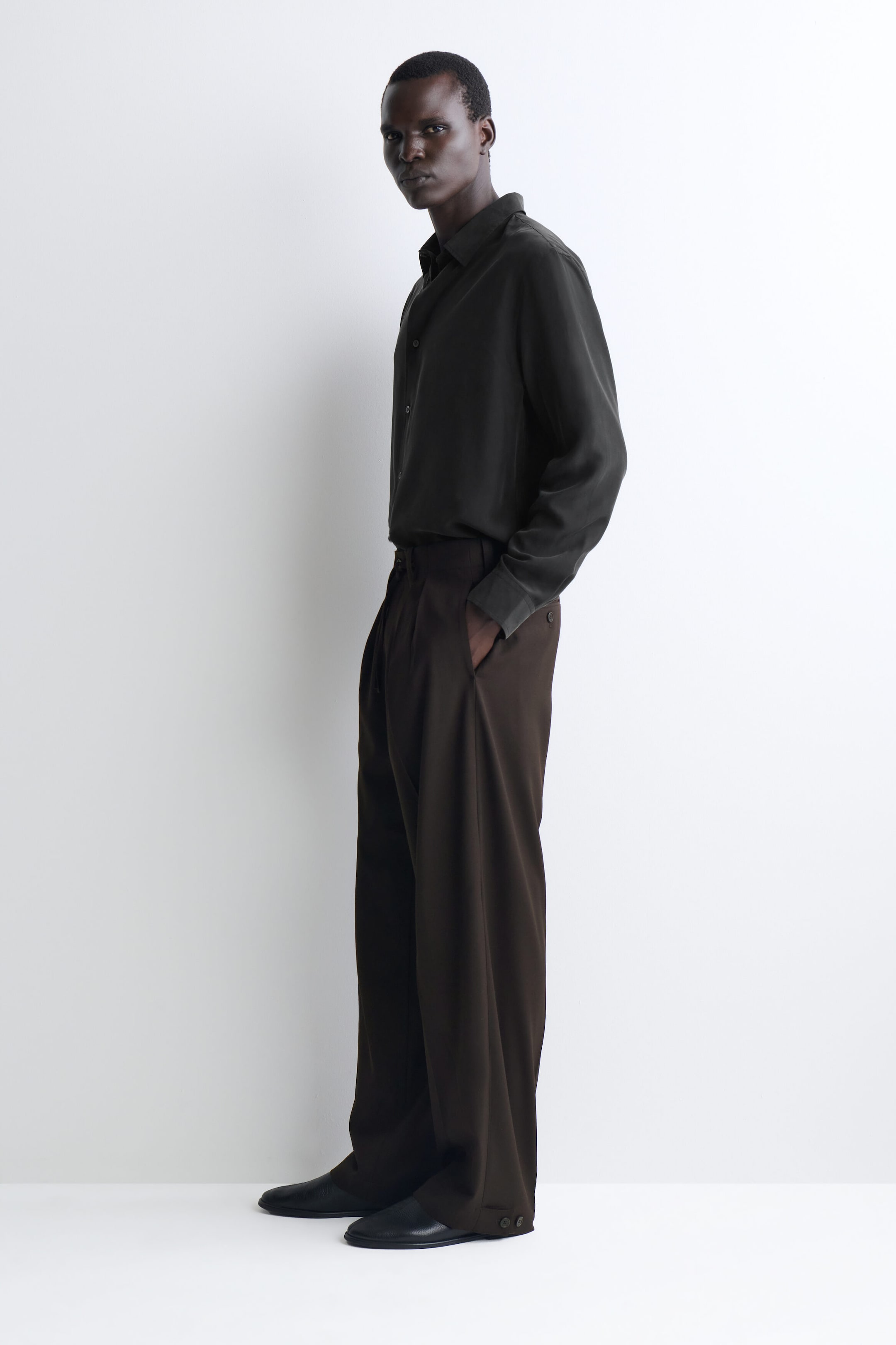 A dark grey long-sleeve button-up shirt is tucked into wide-leg, relaxed-fit dark brown trousers, with the outfit facing left to highlight the fluid drape of the fabrics. The trousers feature subtle button details at the bottom hem and are paired with minimalist black leather slip-on shoes. A dark grey long-sleeve button-up shirt is tucked into wide-leg, relaxed-fit dark brown trousers, with the outfit facing left to highlight the fluid drape of the fabrics. The trousers feature subtle button details at the bottom hem and are paired with minimalist black leather slip-on shoes.