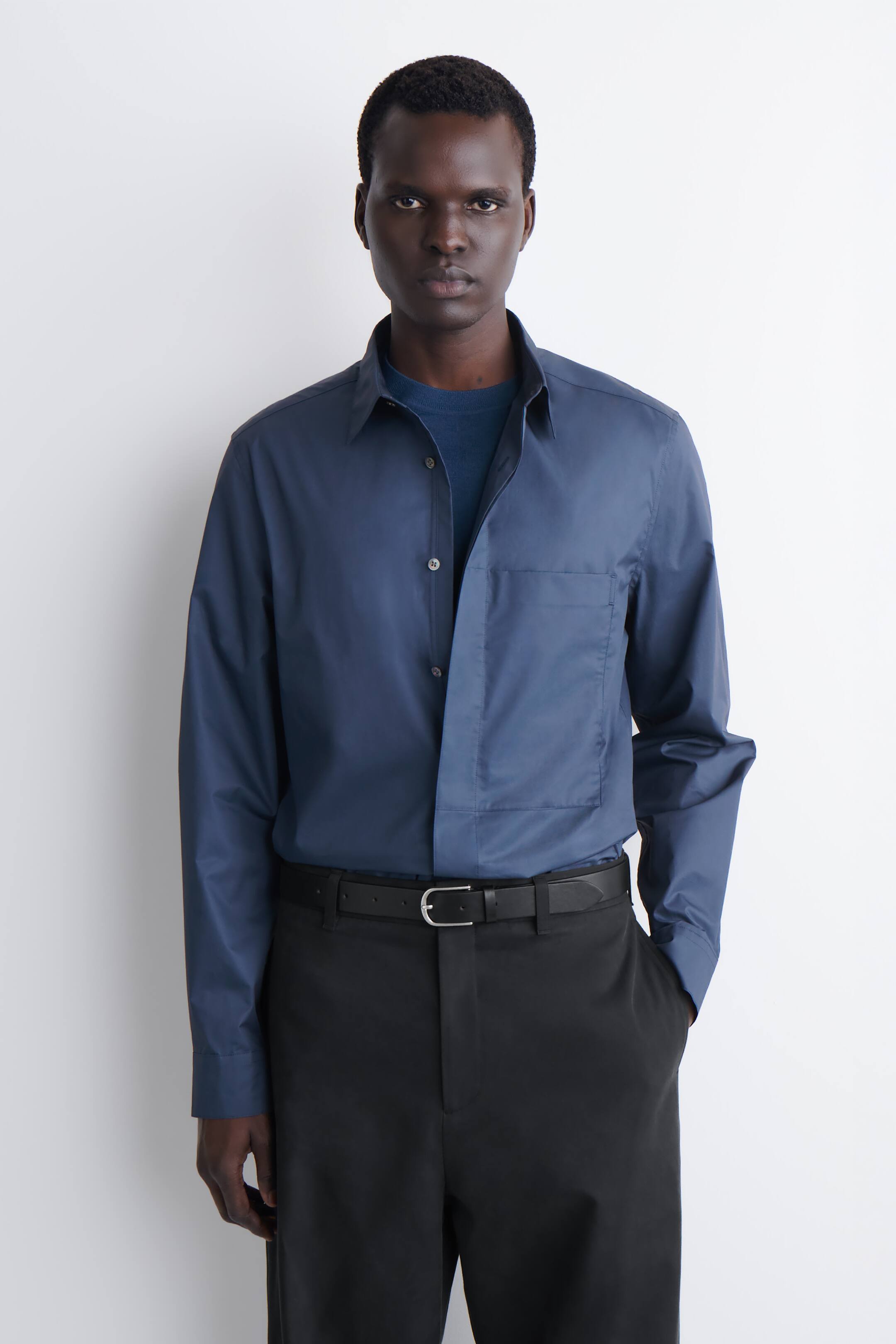 Größeres Bild anzeigen: Ein dunkelblaues langärmeliges Hemd mit Kragen, das eine einzelne Brusttasche und eine teilweise geöffnete Knopfleiste aufweist, wird über einem dunkelblauen Rundhalsshirt präsentiert und mit schwarzen Hosen sowie einem schwarzen Ledergürtel mit silberfarbener Schnalle getragen.