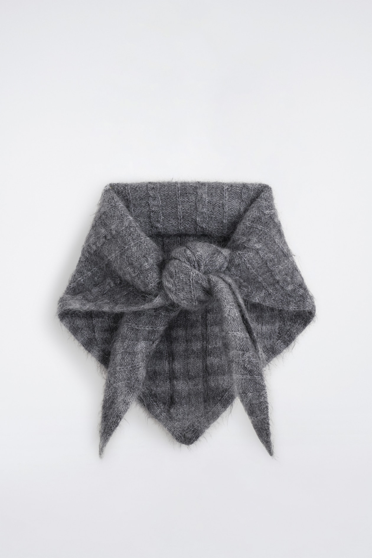 A dark grey checked cable-knit triangle scarf is shown from the front, featuring a fuzzy, brushed knit texture with subtle vertical cable details and checked patterns. The scarf is tied in a loose knot at the center, with its tapered side ends and triangular tip draping downward.