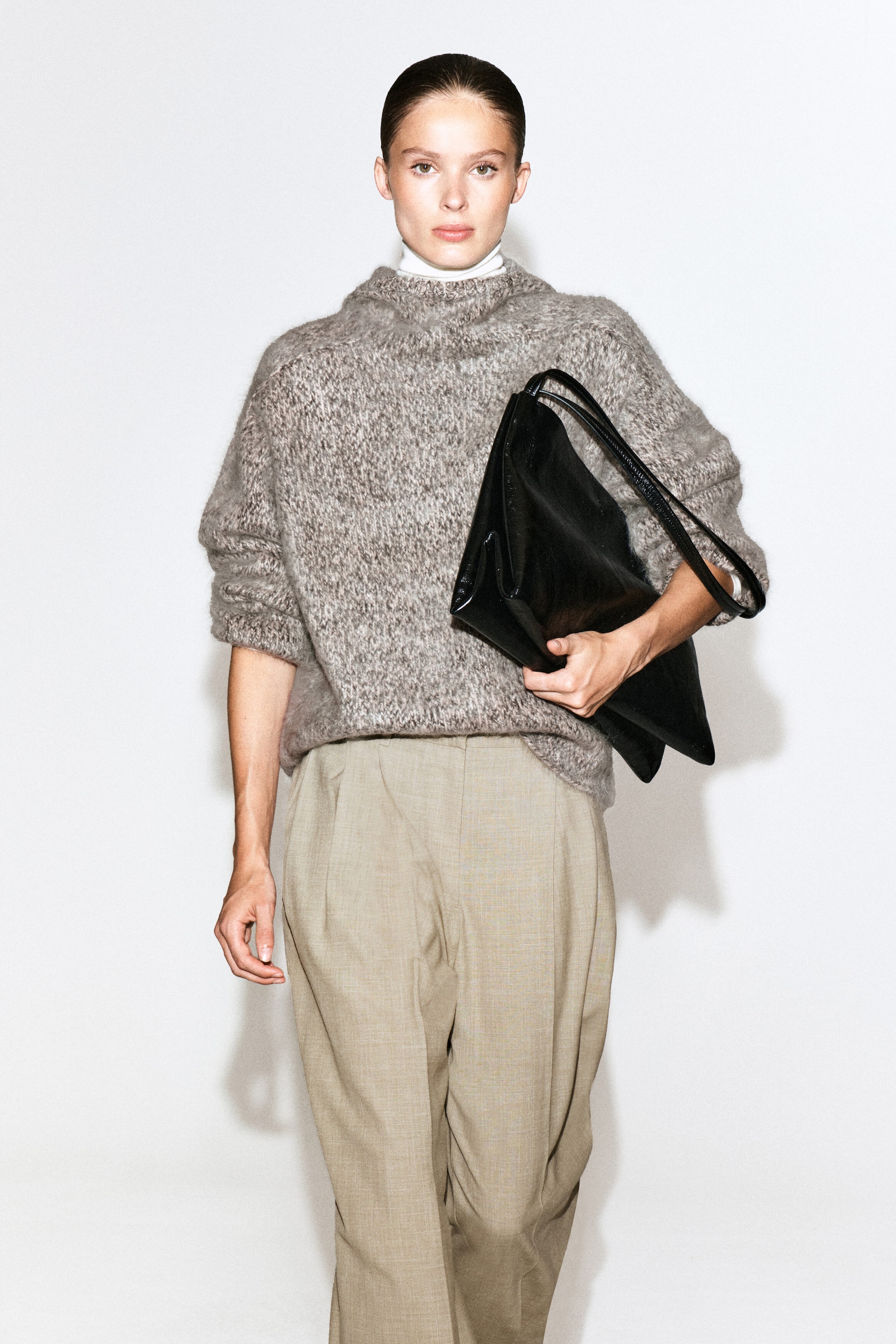 A chunky brushed-cashmere hoodie in a mottled grey-brown textured knit is being worn by the model over a white high-neck top. The relaxed-fit knitwear is styled with beige pleated trousers, with a large black leather bag being carried under one arm. A chunky brushed-cashmere hoodie in a mottled grey-brown textured knit is being worn by the model over a white high-neck top. The relaxed-fit knitwear is styled with beige pleated trousers, with a large black leather bag being carried under one arm.