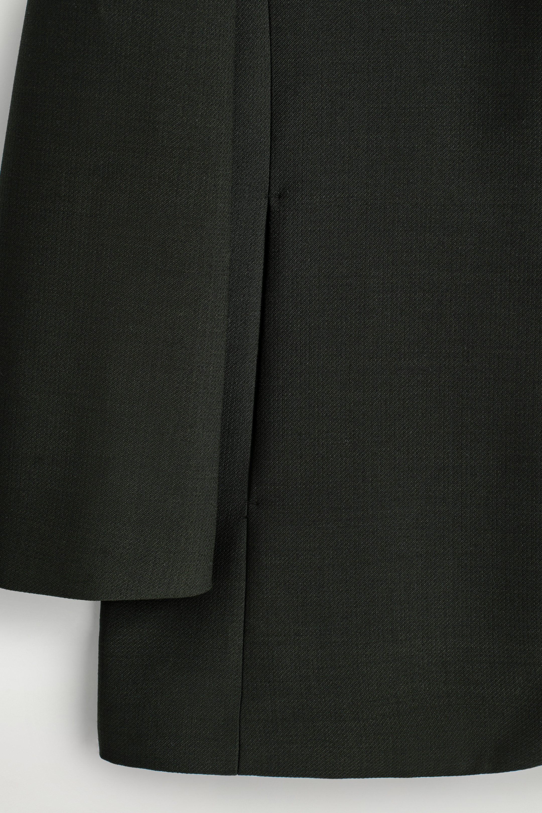 Grösseres Bild ansehen: Eine Nahaufnahme zeigt einen blazer Stoff in tiefem Grün mit einer feinen gewebten Textur. Eine vertikale Naht verläuft in der Mitte, und eine gefaltete Kante desselben Stoffes ist auf der linken Seite sichtbar.