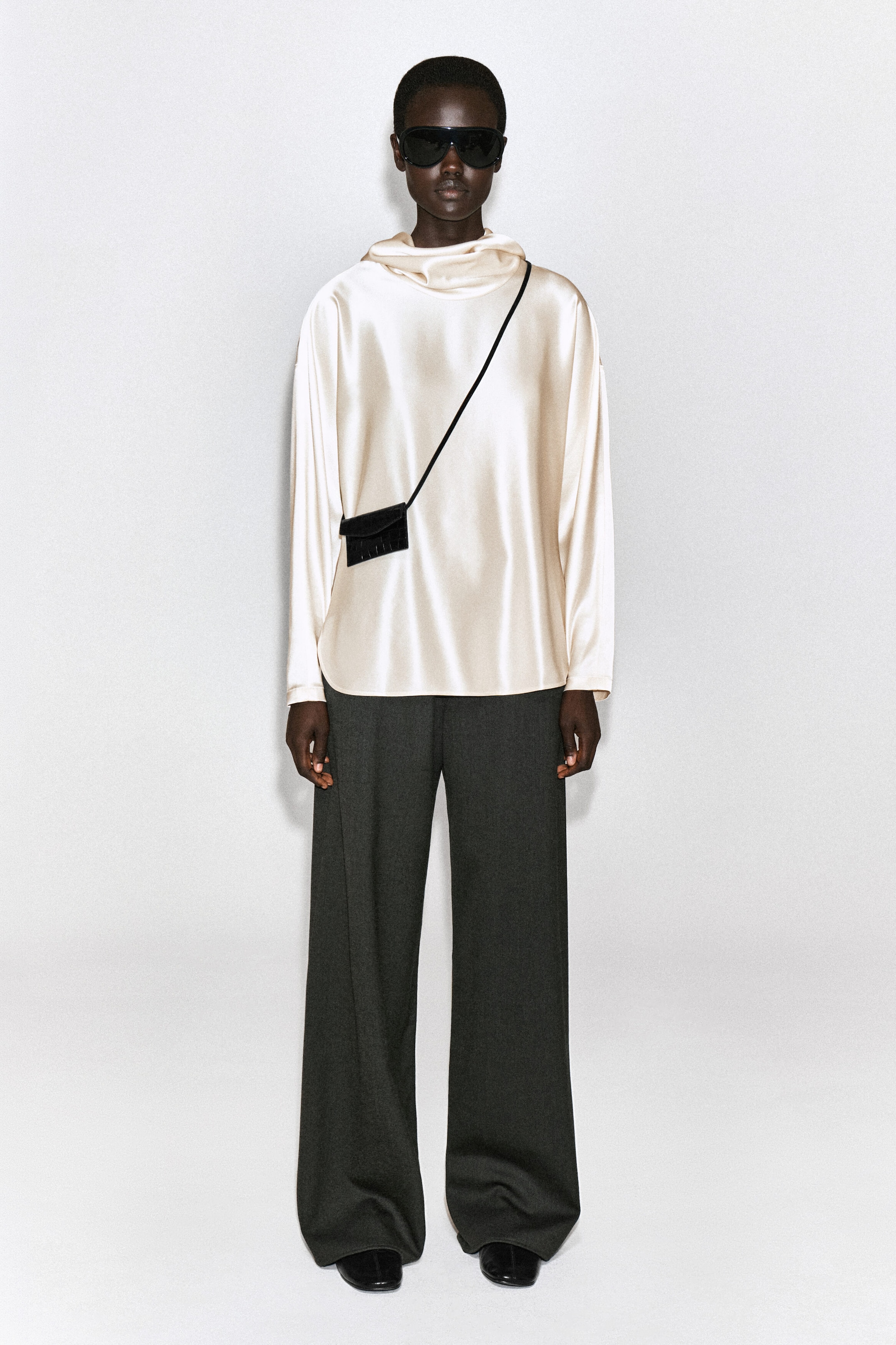 A black leather card holder with a croc-embossed texture and front flap closure is worn crossbody with a thin black strap. The card holder is shown from the front, resting against a cream-colored satin top to highlight its scale and design. A black leather card holder with a croc-embossed texture and front flap closure is worn crossbody with a thin black strap. The card holder is shown from the front, resting against a cream-colored satin top to highlight its scale and design.