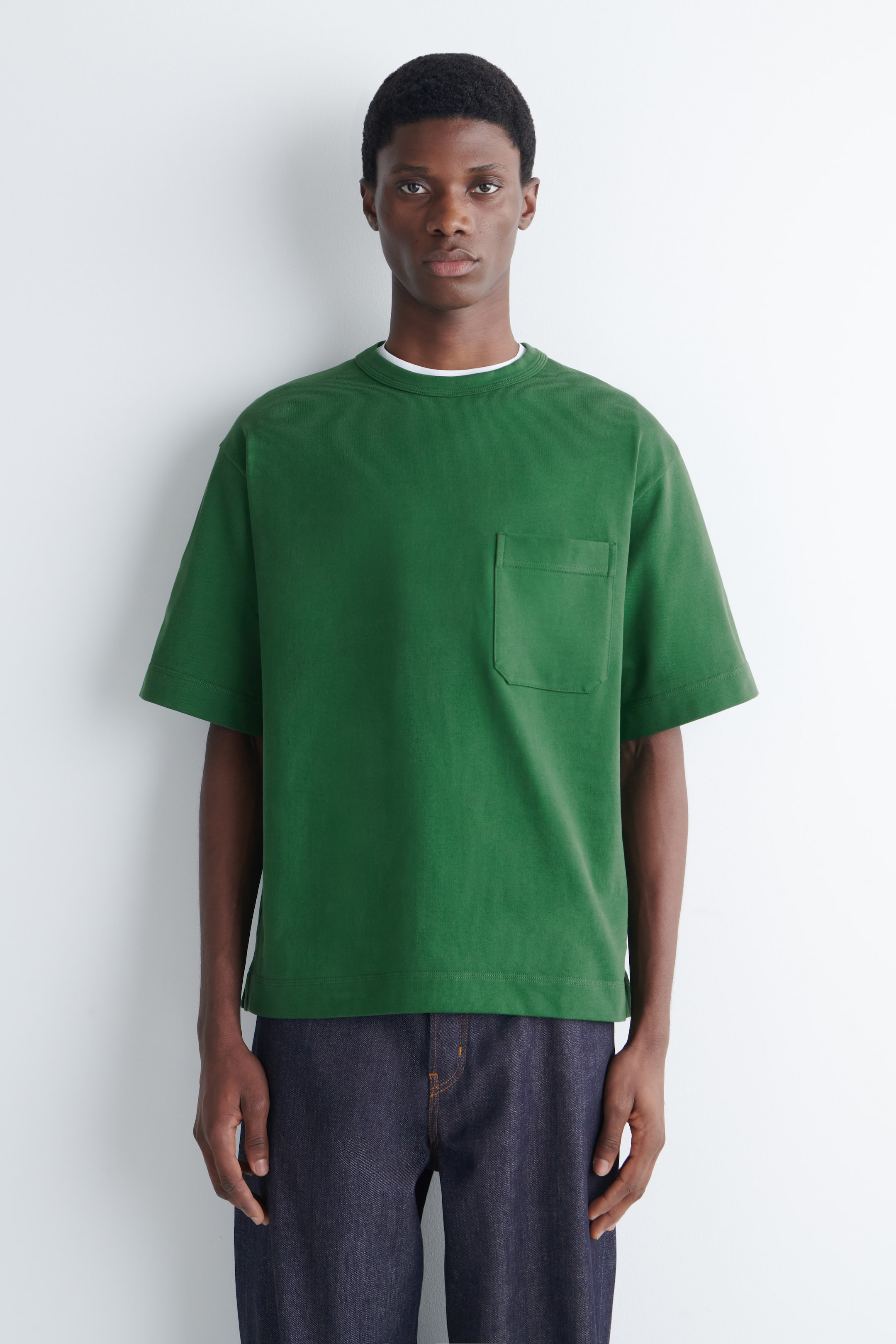 Vis større bilde: Modellen har på seg en oversized grønn T-skjorte med en brystlomme. En hvit rund hals er synlig under, og T-skjorten er kombinert med mørkvaskede denimjeans.