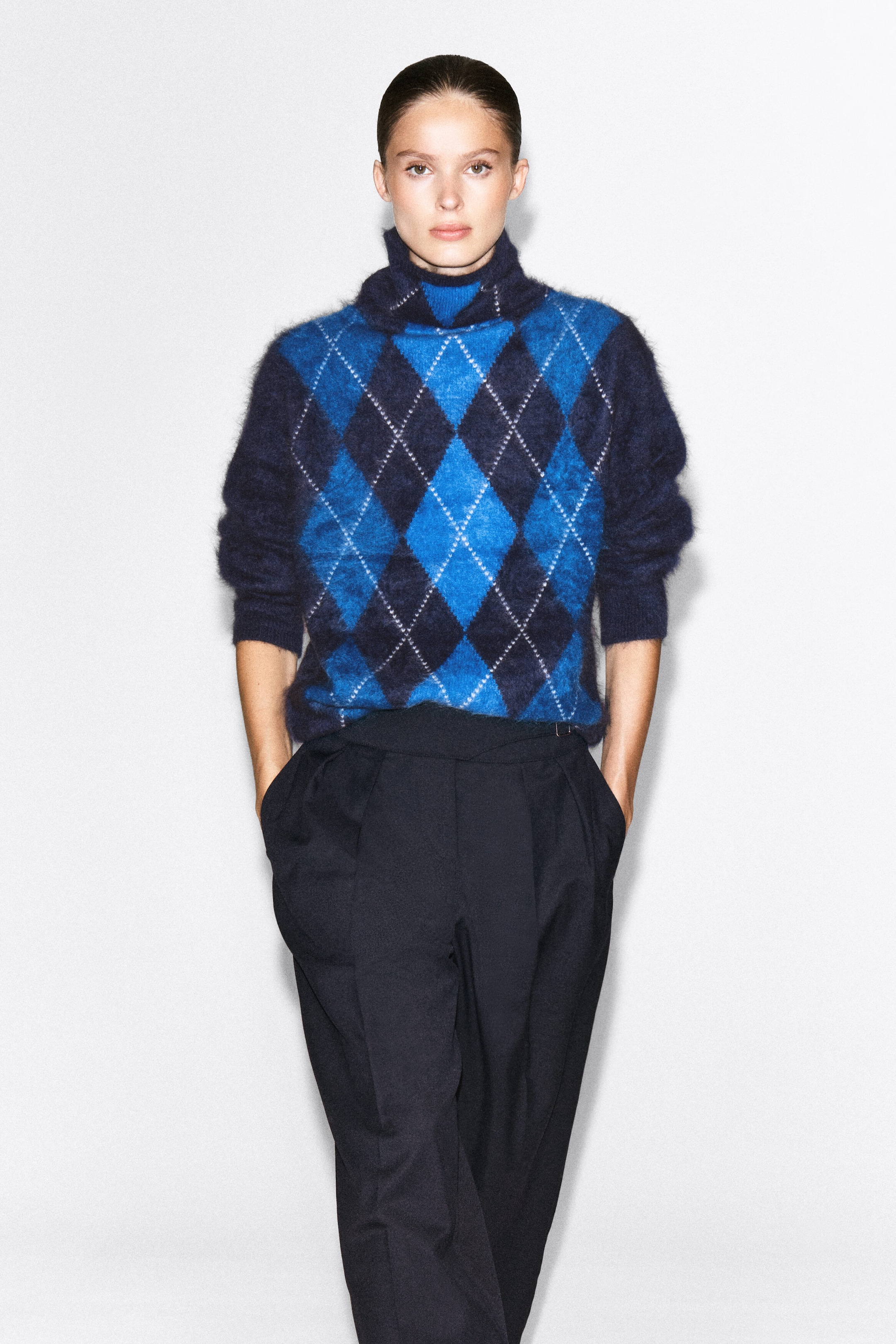 A brushed cashmere jumper in an argyle design, featuring a blue and navy diamond pattern with a fuzzy texture and a high funnel neck, is being worn by the model. The jumper is loosely tucked into dark, high-waisted pleated trousers. A brushed cashmere jumper in an argyle design, featuring a blue and navy diamond pattern with a fuzzy texture and a high funnel neck, is being worn by the model. The jumper is loosely tucked into dark, high-waisted pleated trousers.