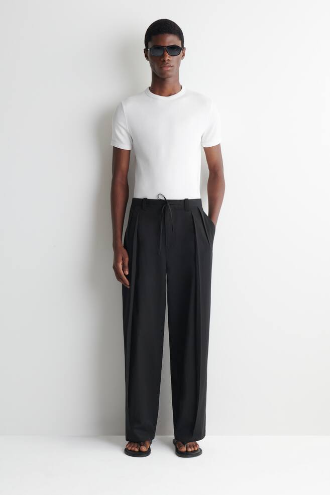 Black trousers are facing forward, featuring a loose, wide-leg fit and a high-waisted design with front pleats. A drawstring is visible at the waist, and side pockets are present, with the trousers pooling at the ankles. - 1