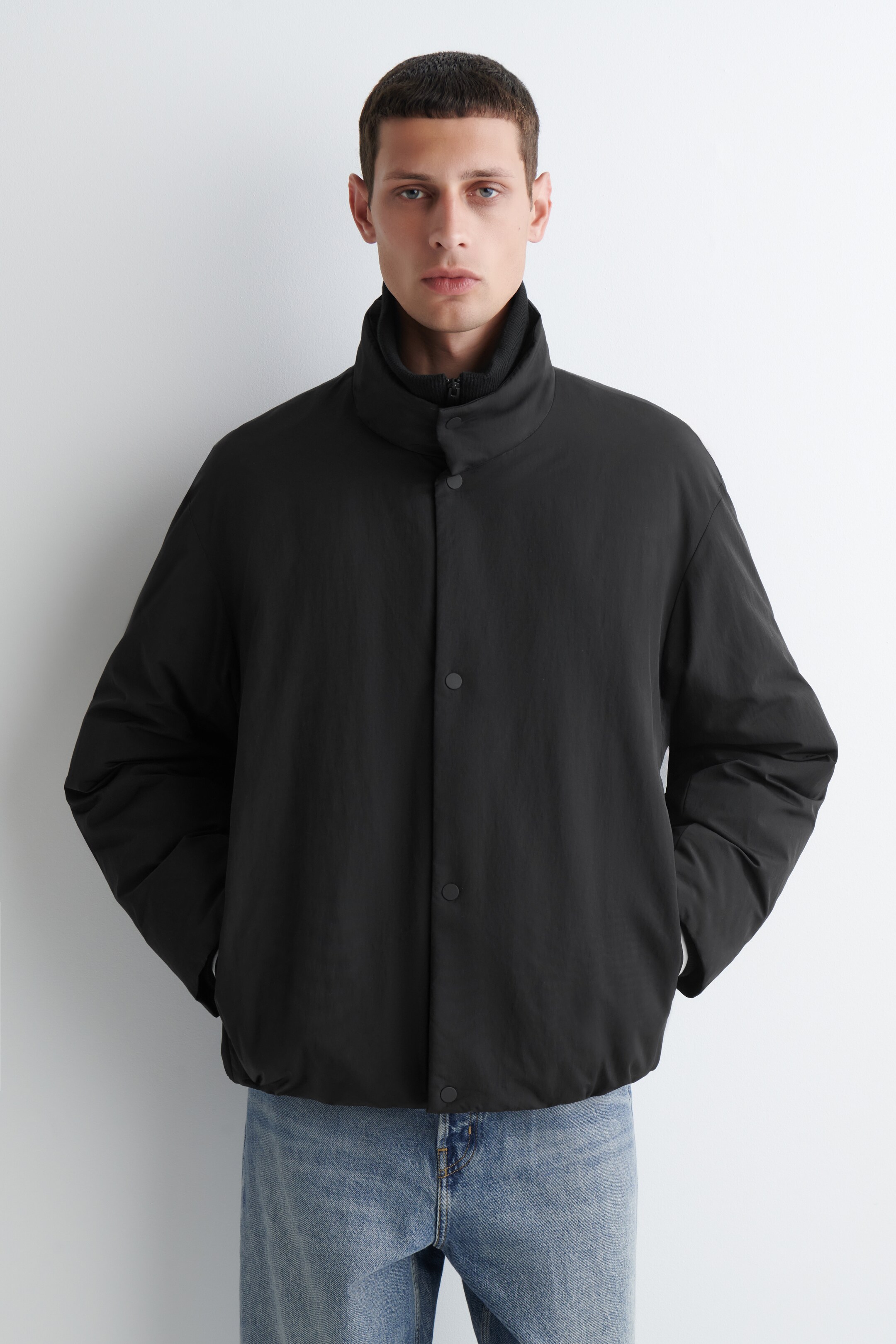 Größeres Bild ansehen: Eine schwarze gesteppte Jacke wird vorwärtsgerichtet präsentiert, mit einem hohen Stehkragen, der durch einen Knopf gesichert ist und eine schwarze gerippte Schicht mit hohem Kragen darunter zeigt. Die Jacke ist mit einer lockeren Passform, einem vollständigen Knopfverschluss vorne und sichtbaren Seitentaschen gestaltet.