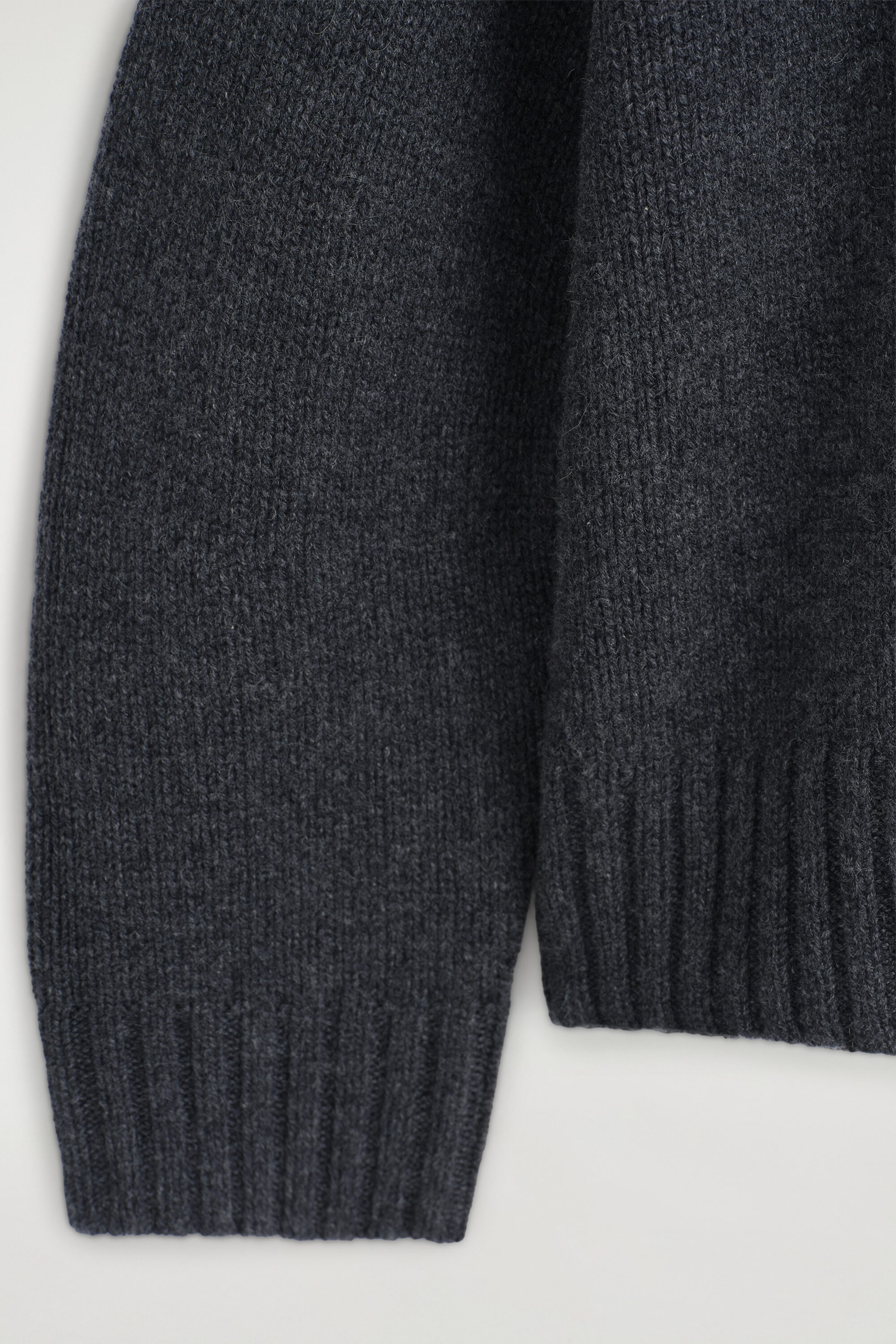 The chunky cashmere funnel-neck jumper is shown in close-up, showcasing its textured dark grey knit fabric. The image focuses on the ribbed detailing of the sleeve cuff and the garment’s hem. The chunky cashmere funnel-neck jumper is shown in close-up, showcasing its textured dark grey knit fabric. The image focuses on the ribbed detailing of the sleeve cuff and the garment’s hem.