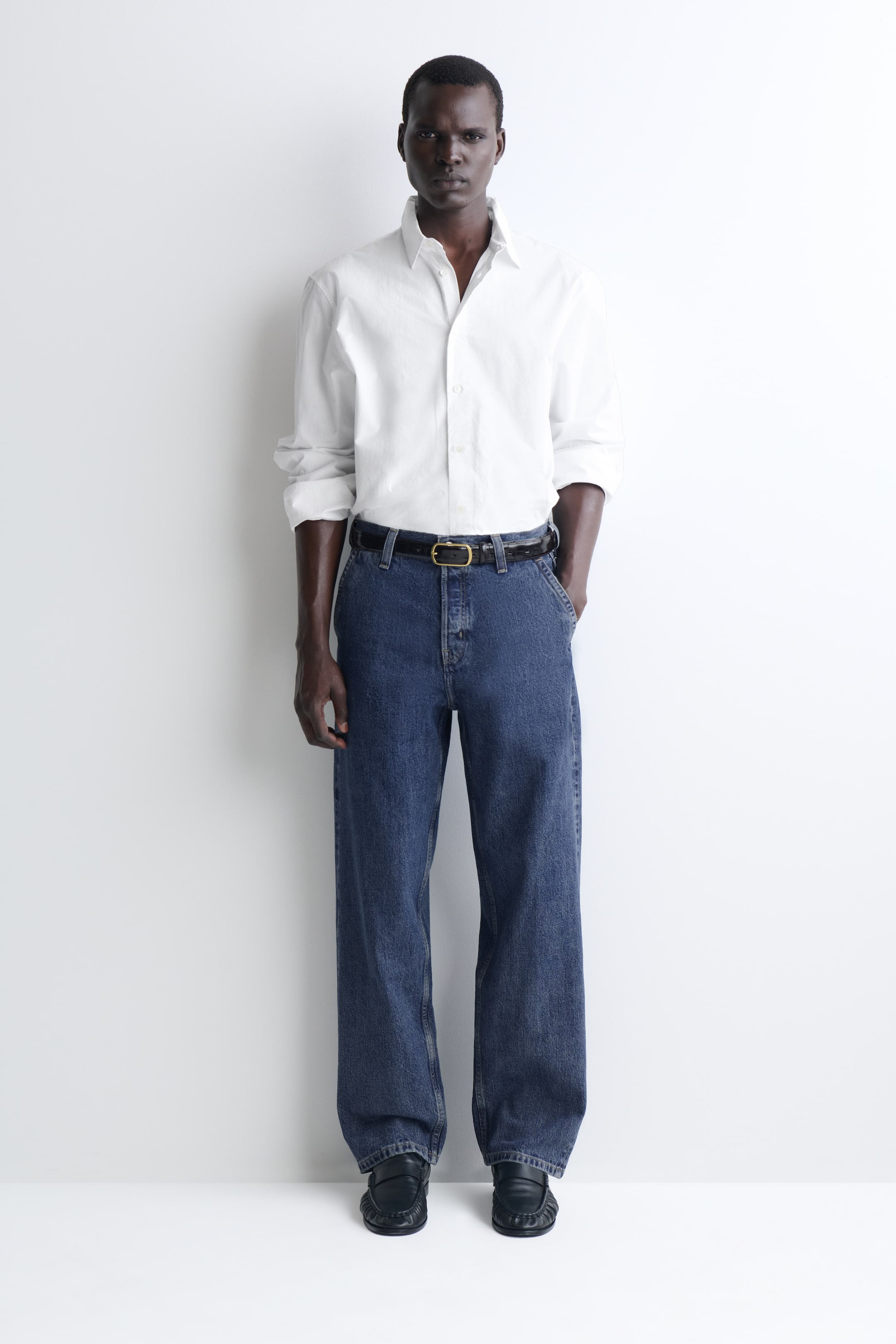 Agrandir l'image: Le pantalon en denim à coupe décontractée et jambe droite, dans un délavage bleu moyen, tombe en une silhouette ample vue de face. Il présente une taille mi-haute, associé à une chemise blanche boutonnée rentrée à l’intérieur et à une ceinture noire, mettant en valeur le design classique à cinq poches et la coupe droite.