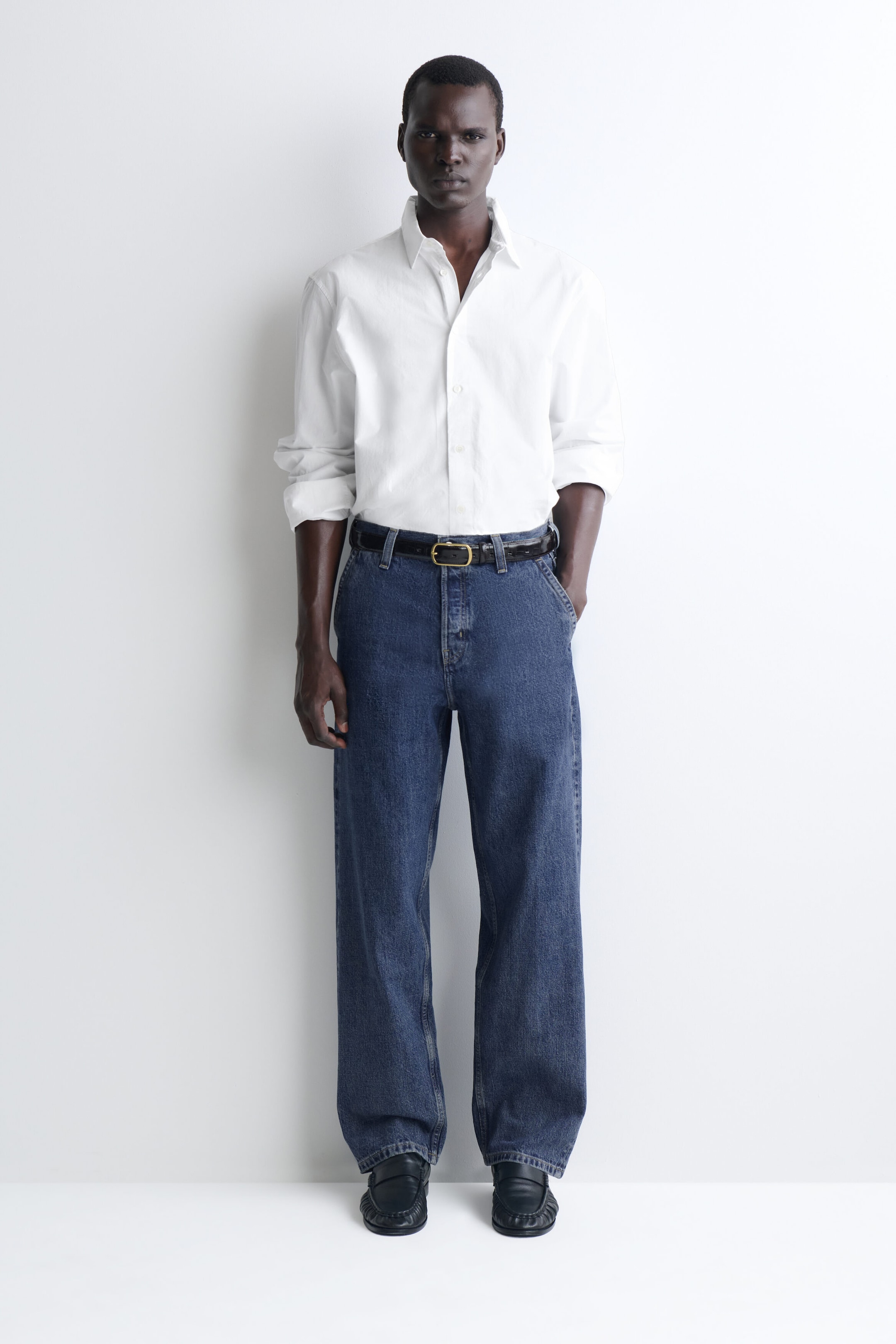 The relaxed-fit straight-leg denim trousers in a medium blue wash are draping in a loose silhouette from a front view. They feature a mid-rise waist styled with a tucked-in white button-up shirt and a black belt, highlighting the classic five-pocket design and straight-leg fit. The relaxed-fit straight-leg denim trousers in a medium blue wash are draping in a loose silhouette from a front view. They feature a mid-rise waist styled with a tucked-in white button-up shirt and a black belt, highlighting the classic five-pocket design and straight-leg fit.