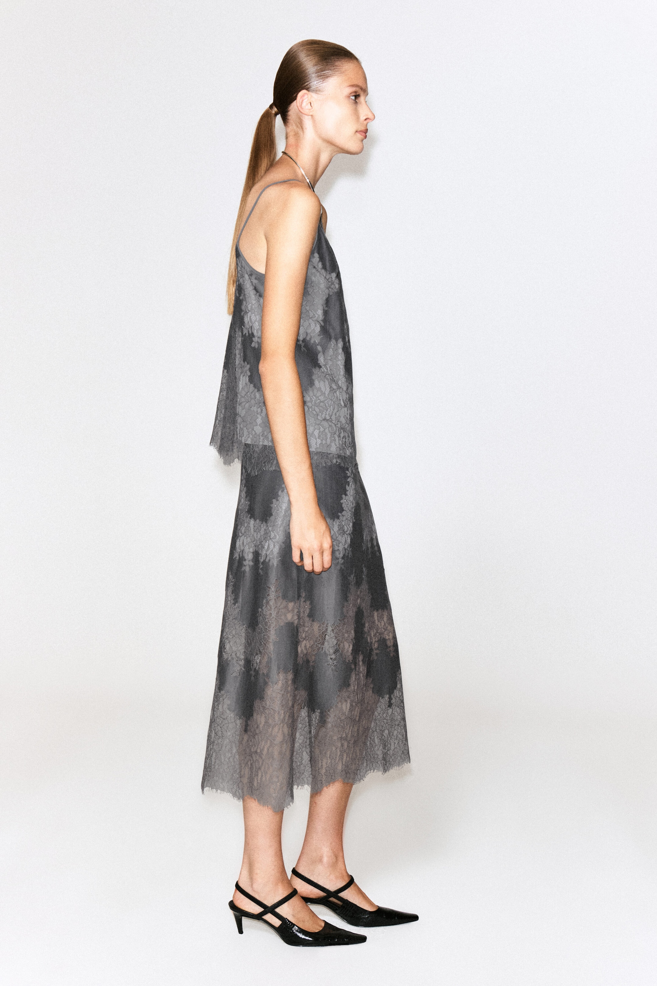 A grey lace-overlay cami top featuring thin spaghetti straps and a semi-sheer patterned lace layer is being worn by the model. The top is paired with a matching lace-overlay midi skirt, shown from a side profile view. A grey lace-overlay cami top featuring thin spaghetti straps and a semi-sheer patterned lace layer is being worn by the model. The top is paired with a matching lace-overlay midi skirt, shown from a side profile view.