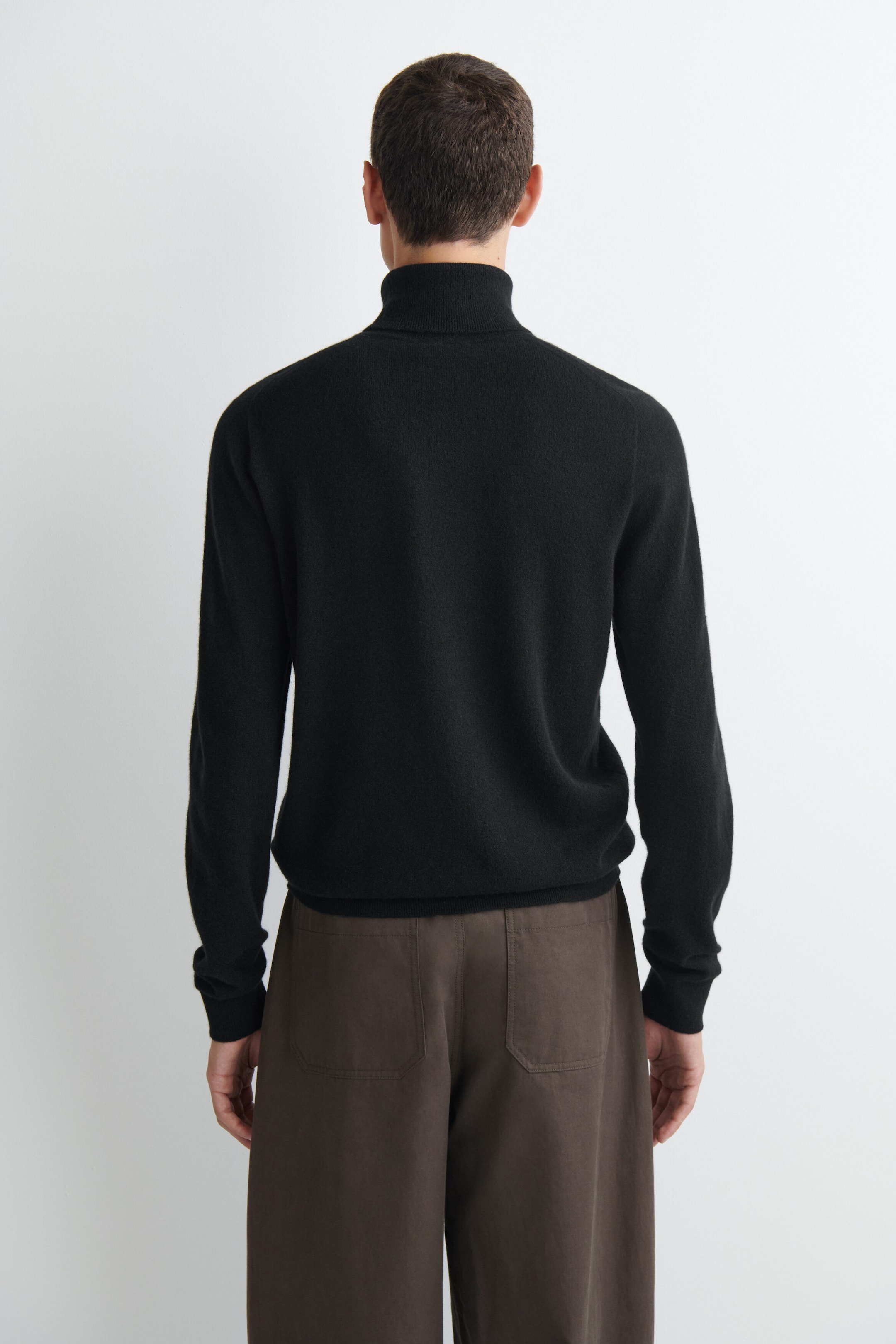 A black knit turtleneck sweater is shown from the back, featuring a ribbed collar, raglan-style sleeves, and ribbed cuffs and hem. The sweater is styled with brown trousers. A black knit turtleneck sweater is shown from the back, featuring a ribbed collar, raglan-style sleeves, and ribbed cuffs and hem. The sweater is styled with brown trousers.