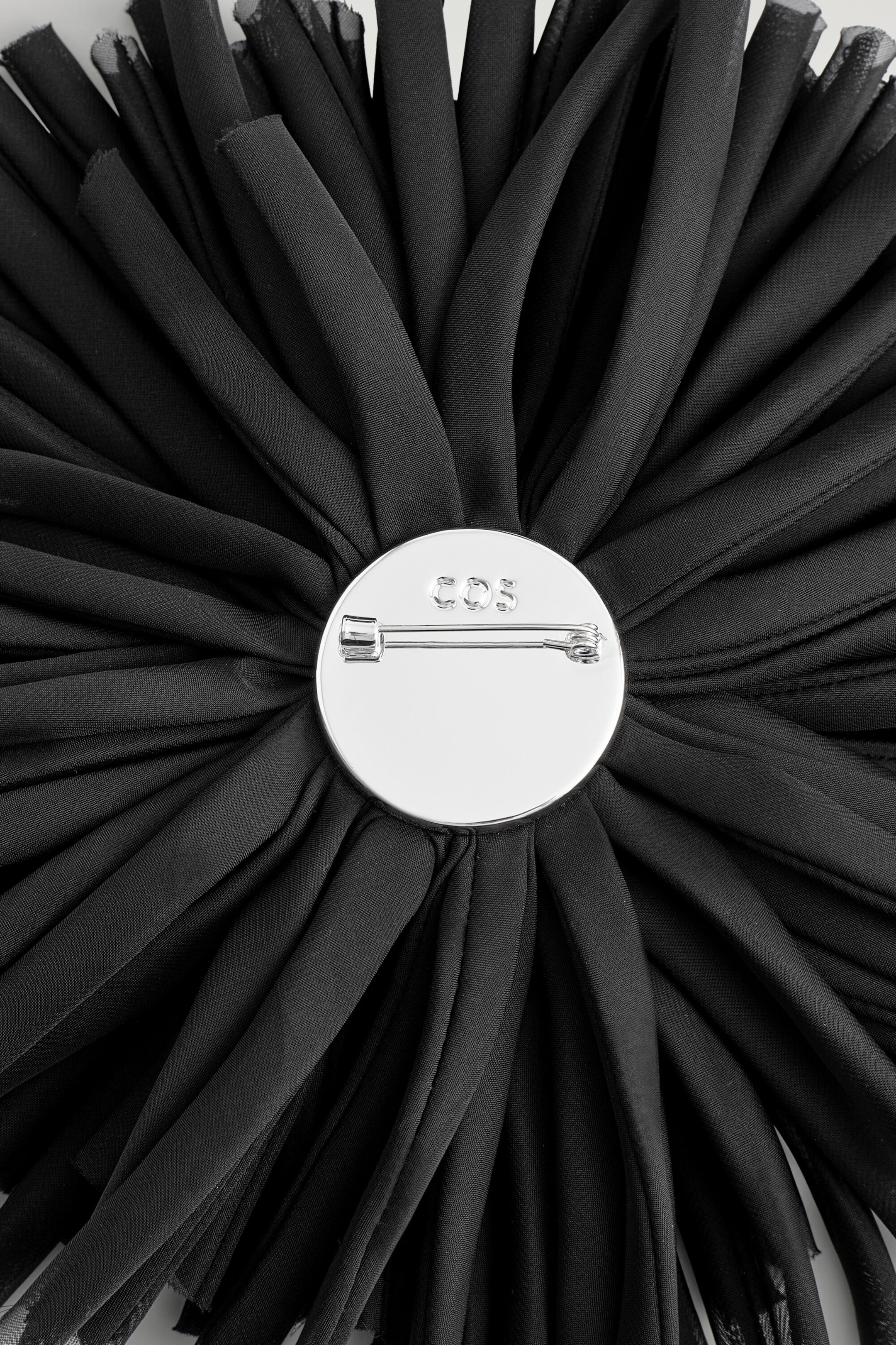 Une broche rosette florale noire est présentée vue de dos, avec une plaque métallique circulaire de teinte argentée polie fixée au centre de pétales en tissu froncés et rayonnants. La plaque métallique affiche une agrafe à épingle horizontale ainsi qu’un logo de marque gravé. Une broche rosette florale noire est présentée vue de dos, avec une plaque métallique circulaire de teinte argentée polie fixée au centre de pétales en tissu froncés et rayonnants. La plaque métallique affiche une agrafe à épingle horizontale ainsi qu’un logo de marque gravé.