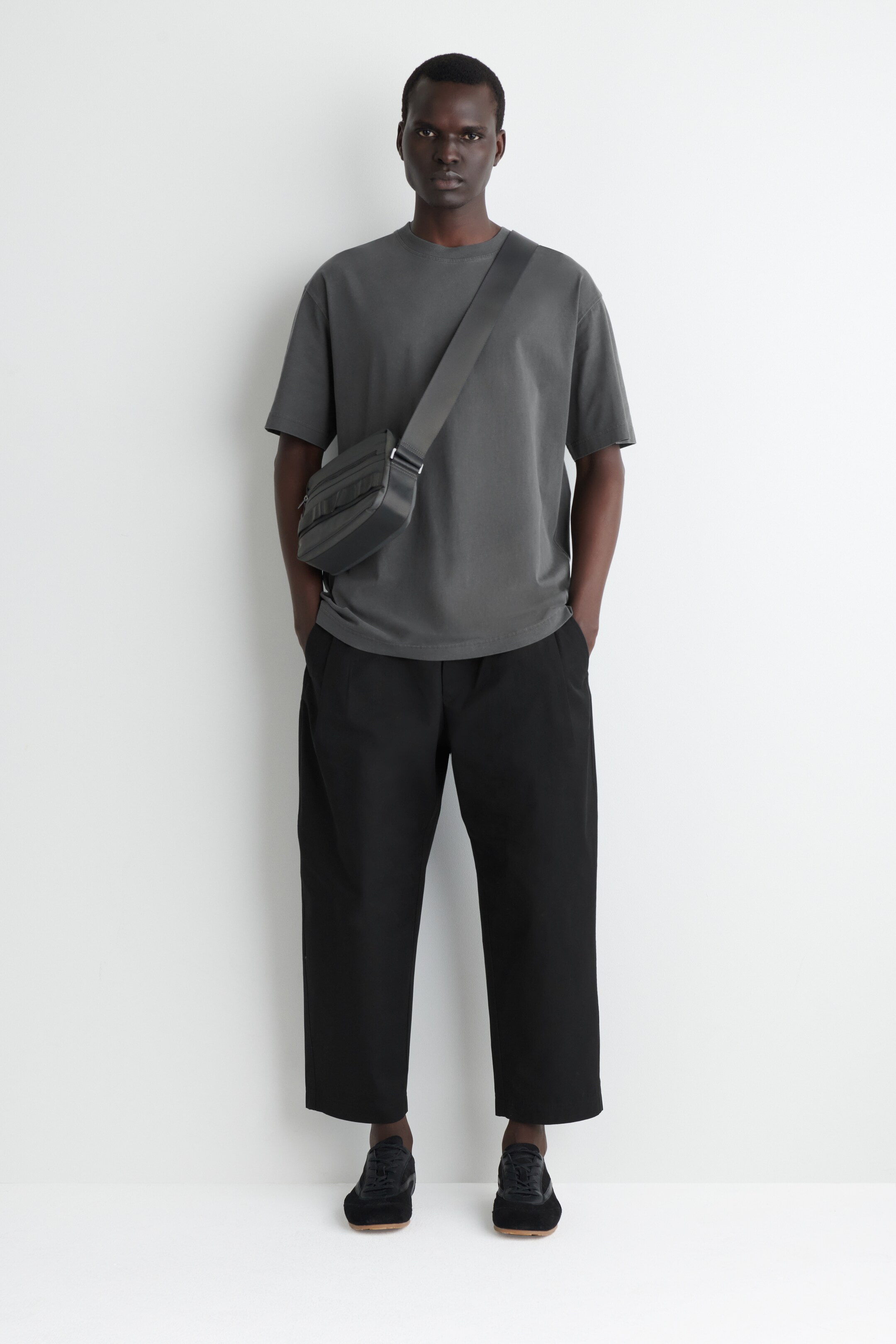 View larger image: A front-facing outfit is being presented, featuring a dark gray oversized crew-neck t-shirt and black wide-leg cropped trousers. A dark gray cross-body bag with multiple zippered compartments is being worn across the chest, complemented by black low-top sneakers with light brown soles.