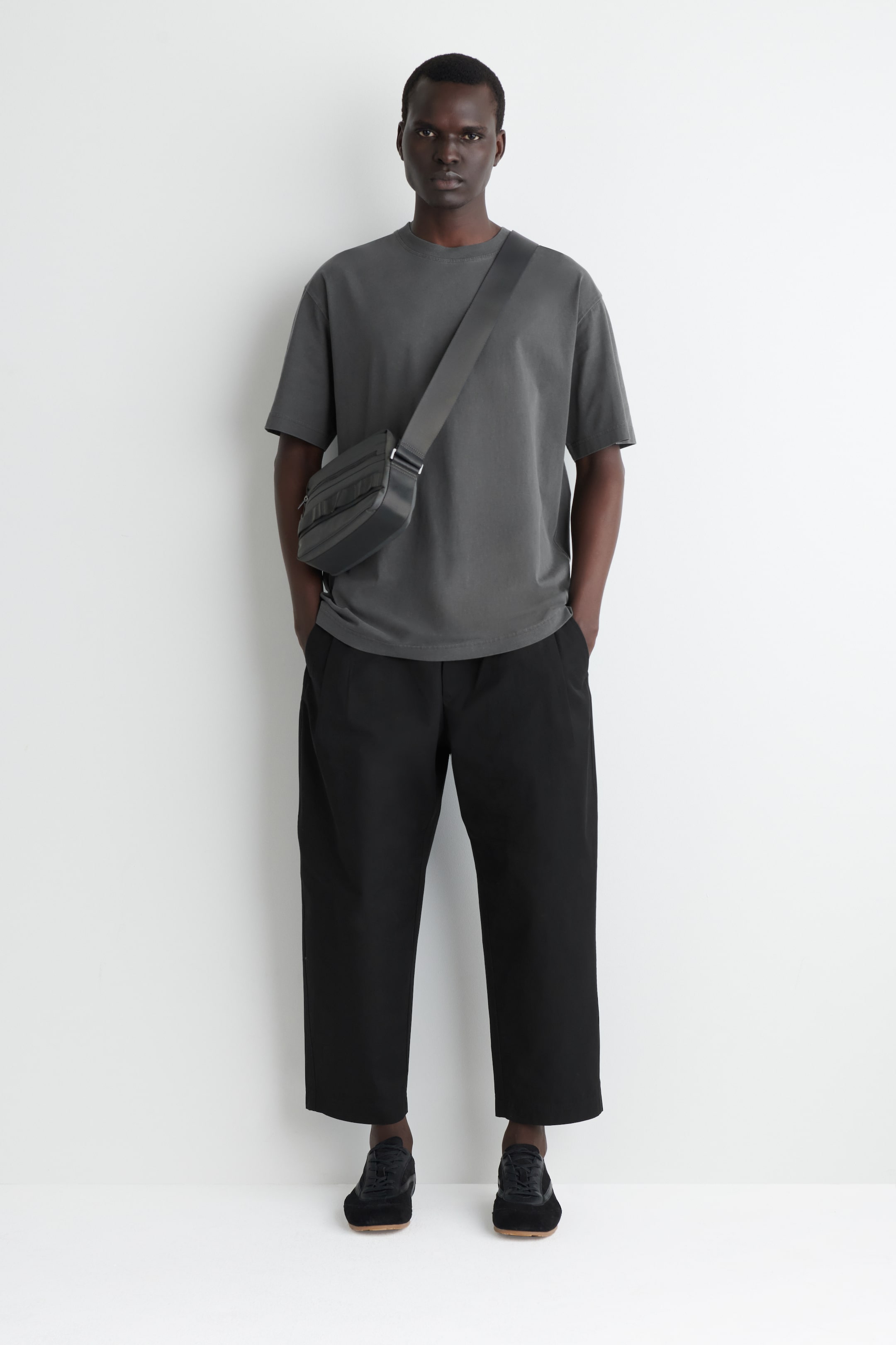 Agrandir l'image: Une tenue orientée vers l'avant est présentée, comprenant un t-shirt oversize à col rond gris foncé et un pantalon large noir à jambes courtes. Un sac bandoulière gris foncé avec plusieurs compartiments zippés est porté en travers de la poitrine, complété par des baskets basses noires avec des semelles marron clair.