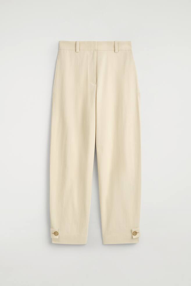 Pantalones de color crema están de frente, con trabillas para el cinturón en la cintura y una costura visible en la bragueta. Las piernas de corte relajado están recortadas, y cada dobladillo está asegurado en el lado exterior con un único botón redondo de color marrón claro. - 1