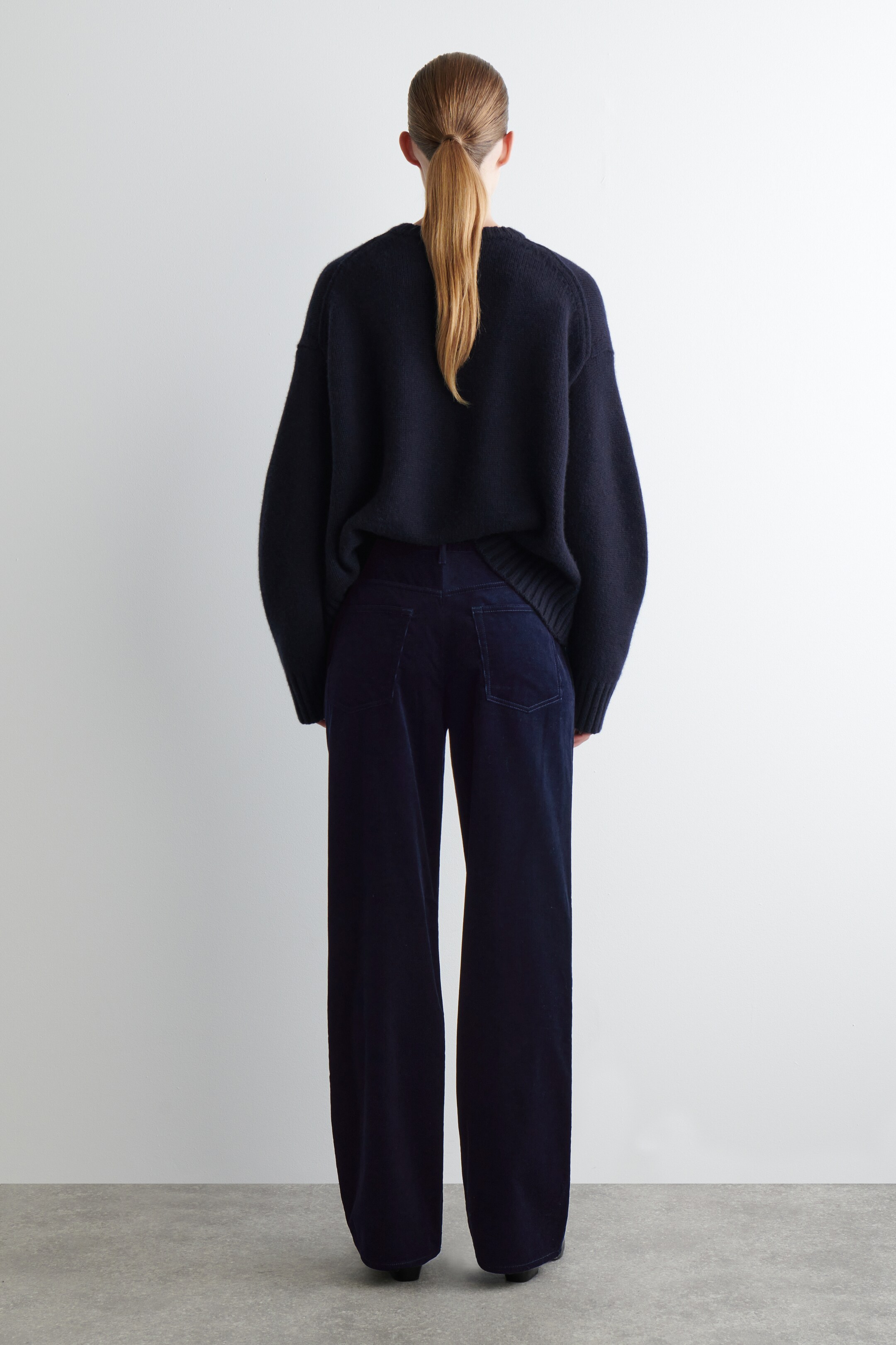 Agrandir l'image: Une personne est debout, tournée dos, portant un pull en tricot bleu foncé à coupe décontractée et un pantalon large assorti. Le pull oversize présente un ourlet côtelé, tandis que le pantalon arbore une texture en velours côtelé doux et des poches arrière plaquées.