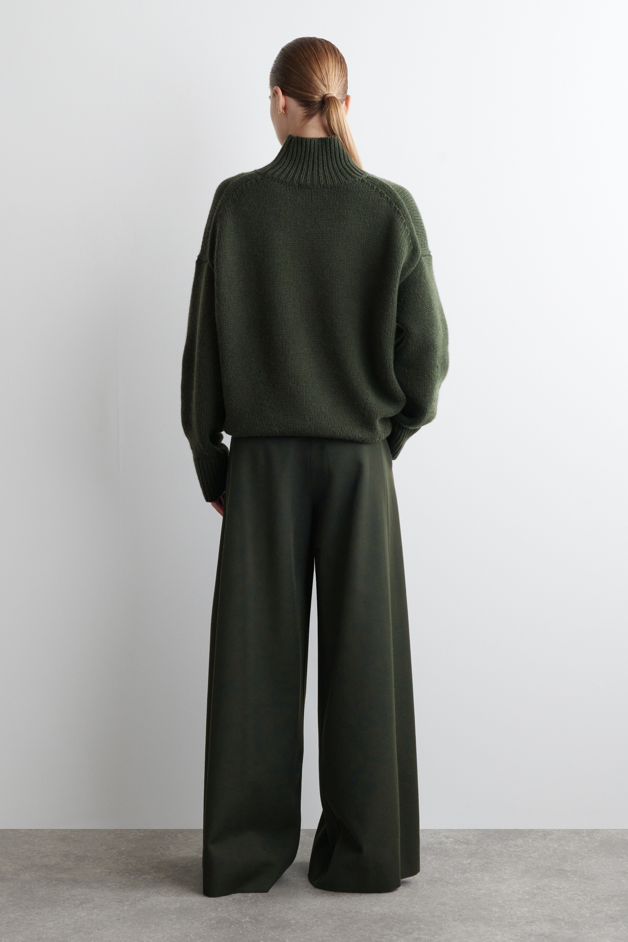 Größeres Bild anzeigen: Ein dunkelgrüner Strickpullover wird von hinten gezeigt, mit einem grob gerippten Trichterkragen, überschnittenen Schultern und vollen, abgerundeten Ärmeln. Er präsentiert eine lockere, übergroße Passform, die locker über den Körper fällt.