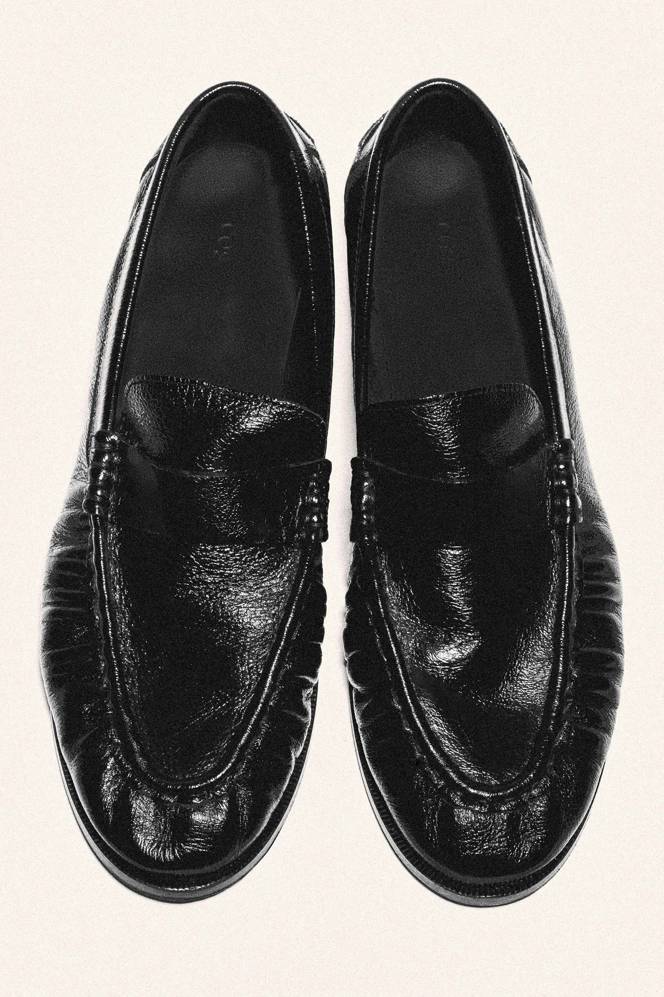 Visualizza immagine più grande: Un paio di mocassini loafer in pelle nera è mostrato da una prospettiva dall’alto, caratterizzato da una texture lucida e increspata e dalle classiche fascette penny sulla tomaia. Il design mette in evidenza una cucitura moc-toe ben visibile con dettagli arricciati attorno alla punta arrotondata.