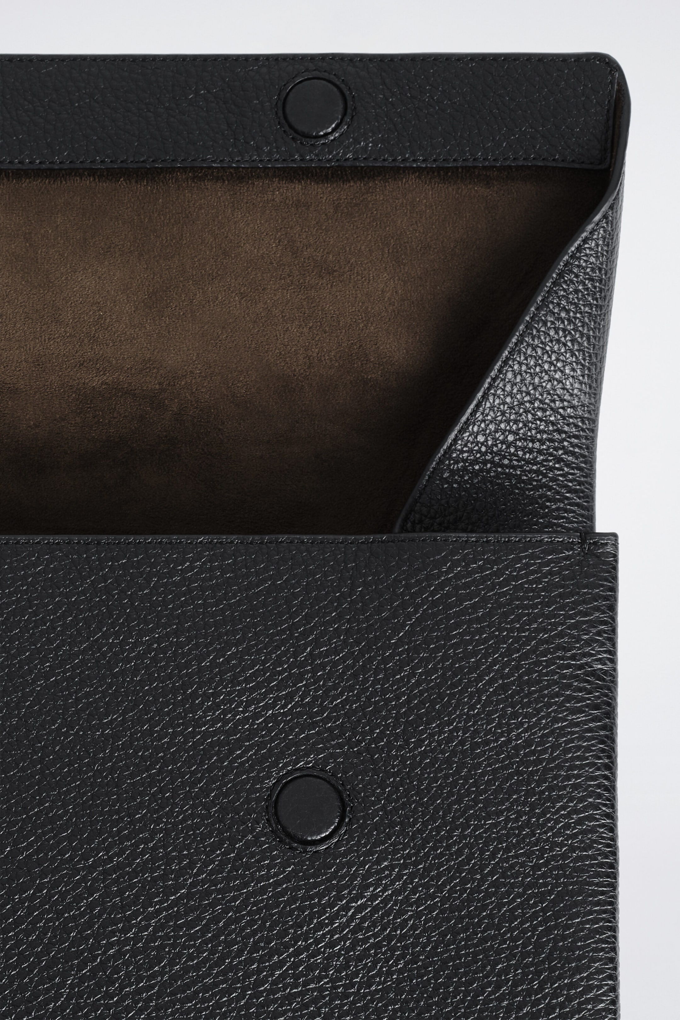 The envelope-style laptop sleeve, featuring a black pebbled leather exterior, is shown with its front flap partially open to reveal a soft brown suede interior lining. Two matching, leather-covered circular magnetic snap closures are visible on the flap and the body of the sleeve. The envelope-style laptop sleeve, featuring a black pebbled leather exterior, is shown with its front flap partially open to reveal a soft brown suede interior lining. Two matching, leather-covered circular magnetic snap closures are visible on the flap and the body of the sleeve.
