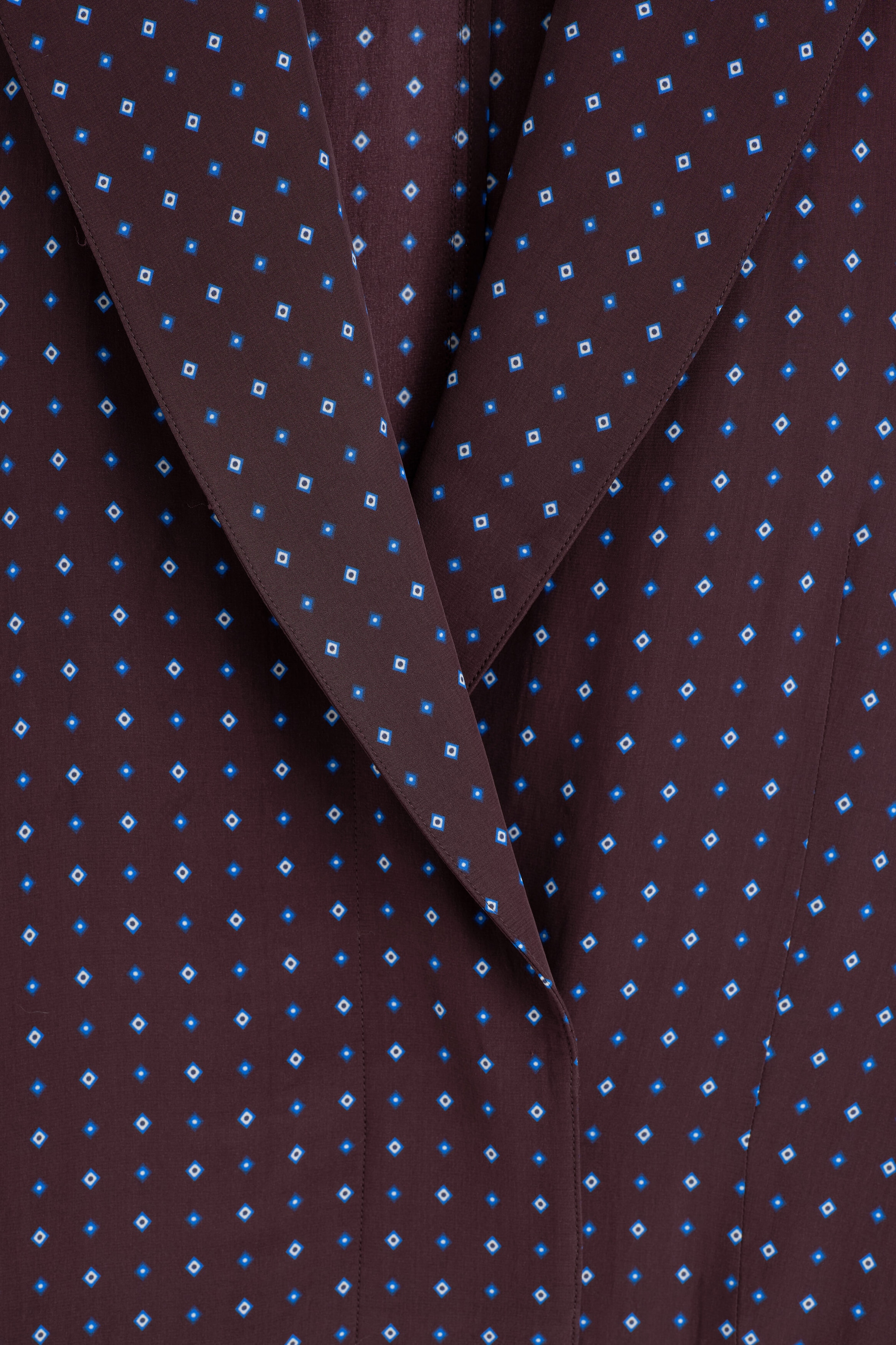 The diamond-print double-breasted blazer is shown in a close-up front view, featuring dark brown fabric patterned with small blue and white geometric diamonds. The design features wide, pointed lapels overlapping at the centre. The diamond-print double-breasted blazer is shown in a close-up front view, featuring dark brown fabric patterned with small blue and white geometric diamonds. The design features wide, pointed lapels overlapping at the centre.